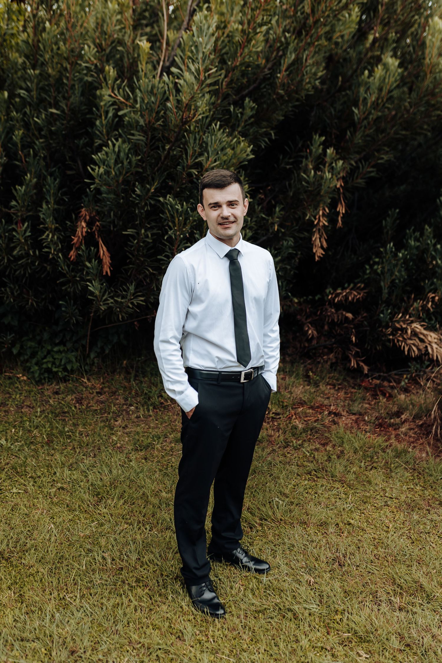 A person in formal business attire poses with hands in pockets against a natural backdrop.