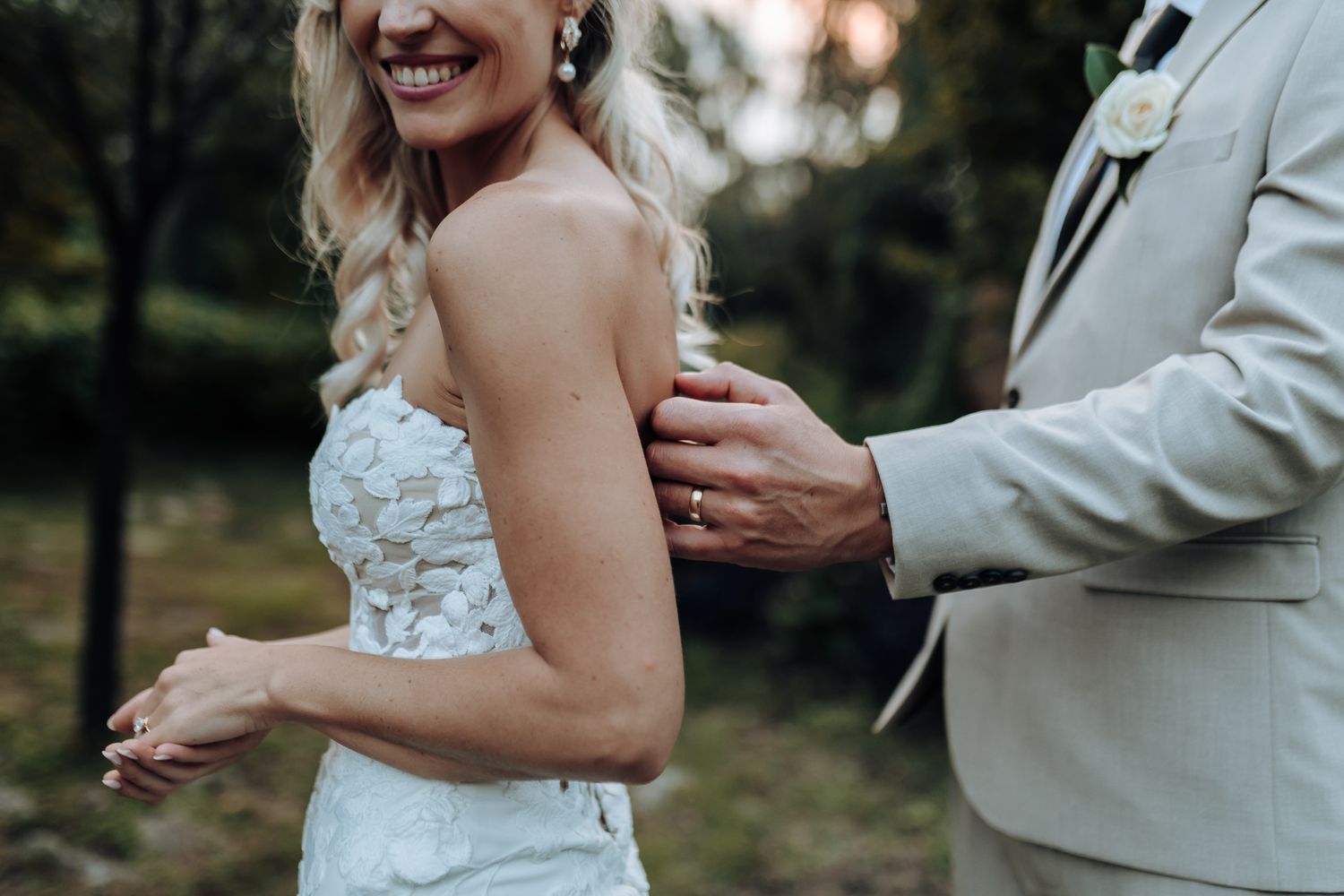 Close-up of a lace wedding dress detail and light beige suit sleeve in an outdoor garden setting.