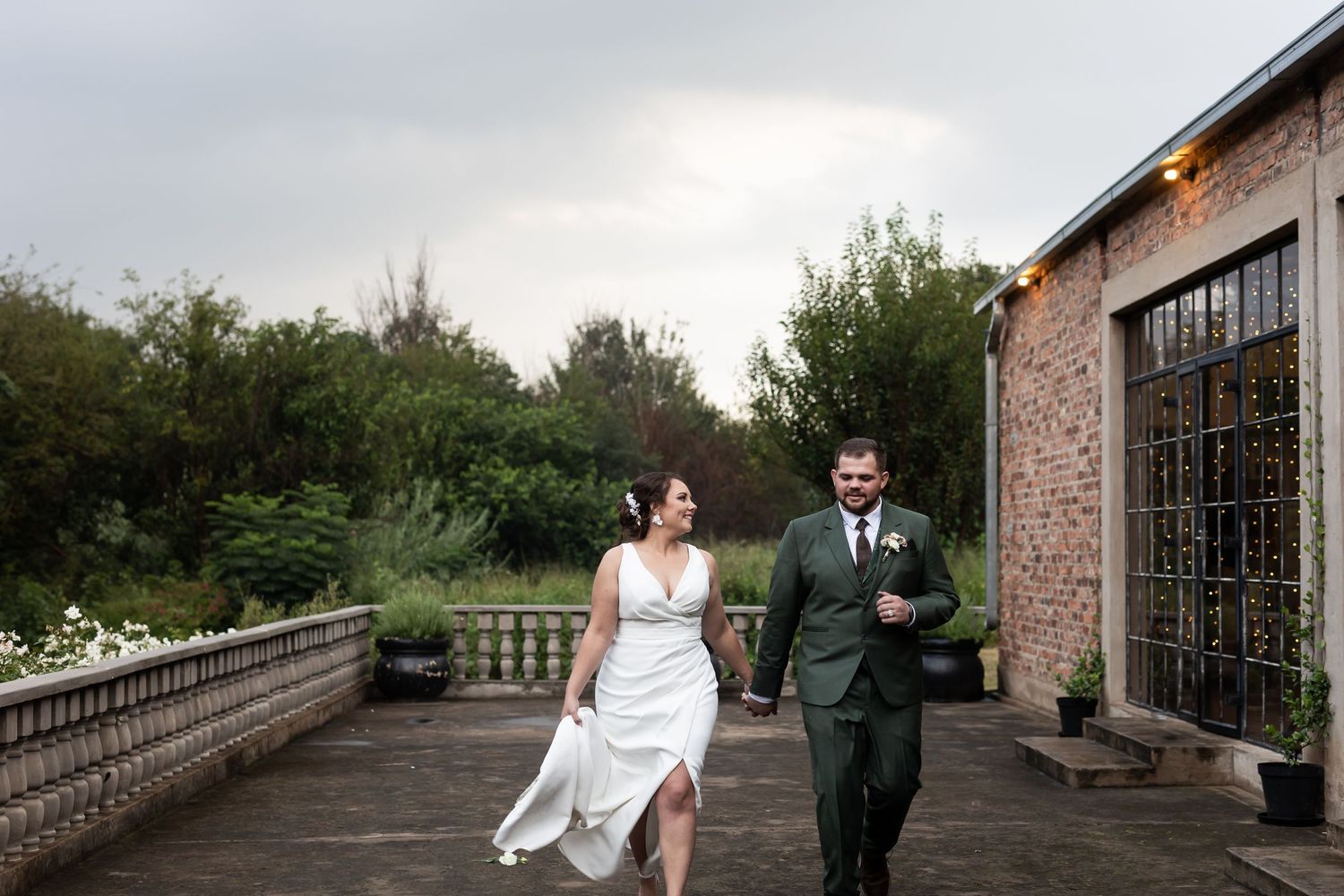 Couple walking together on a stone terrace with greenery in the background and string lights overhead.