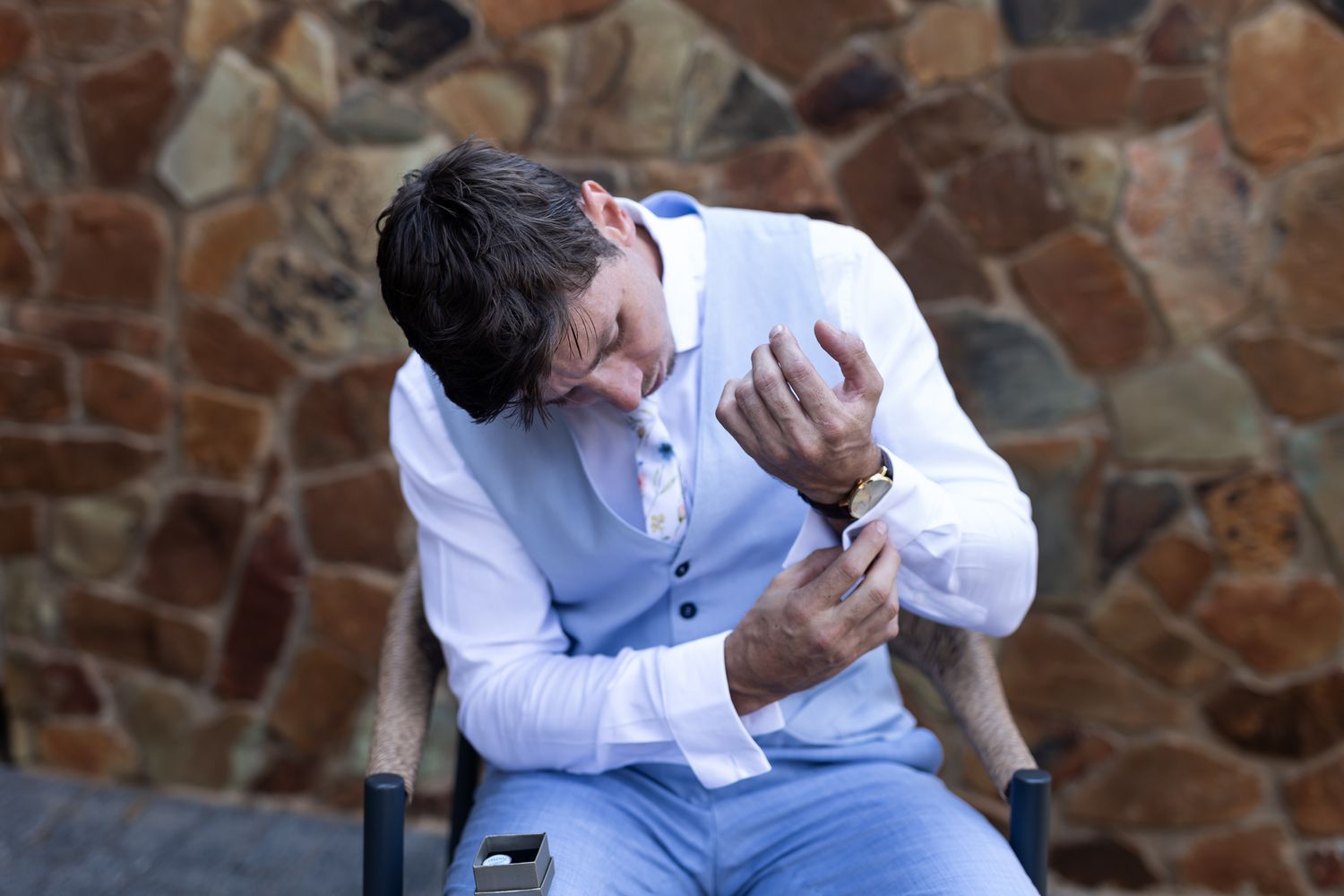 Person in light blue vest and white shirt sits emotively in front of stone wall.