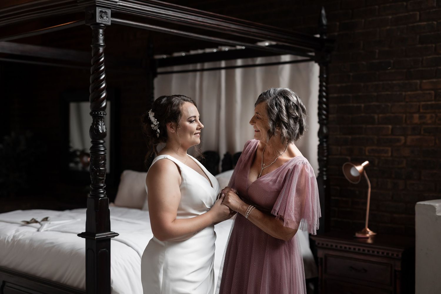 Two people share an intimate moment in a bedroom with dark wooden furniture and brick walls.