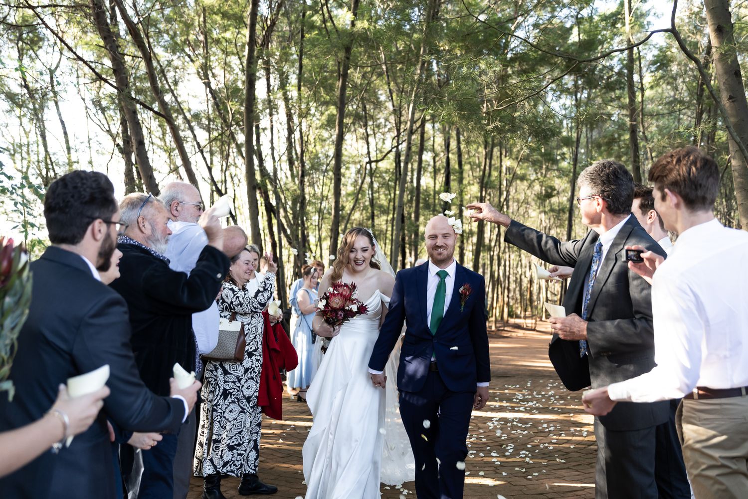 Wedding celebration with guests throwing confetti in a sun-dappled forest setting.