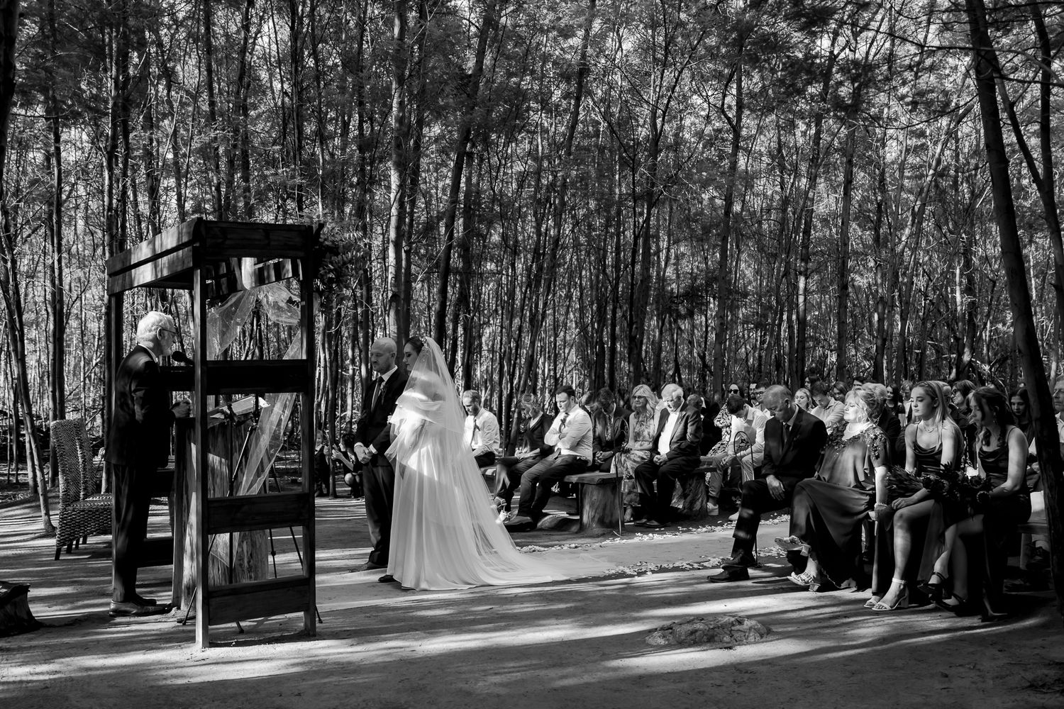 A black and white photograph of a forest wedding ceremony with wooden benches and guests.