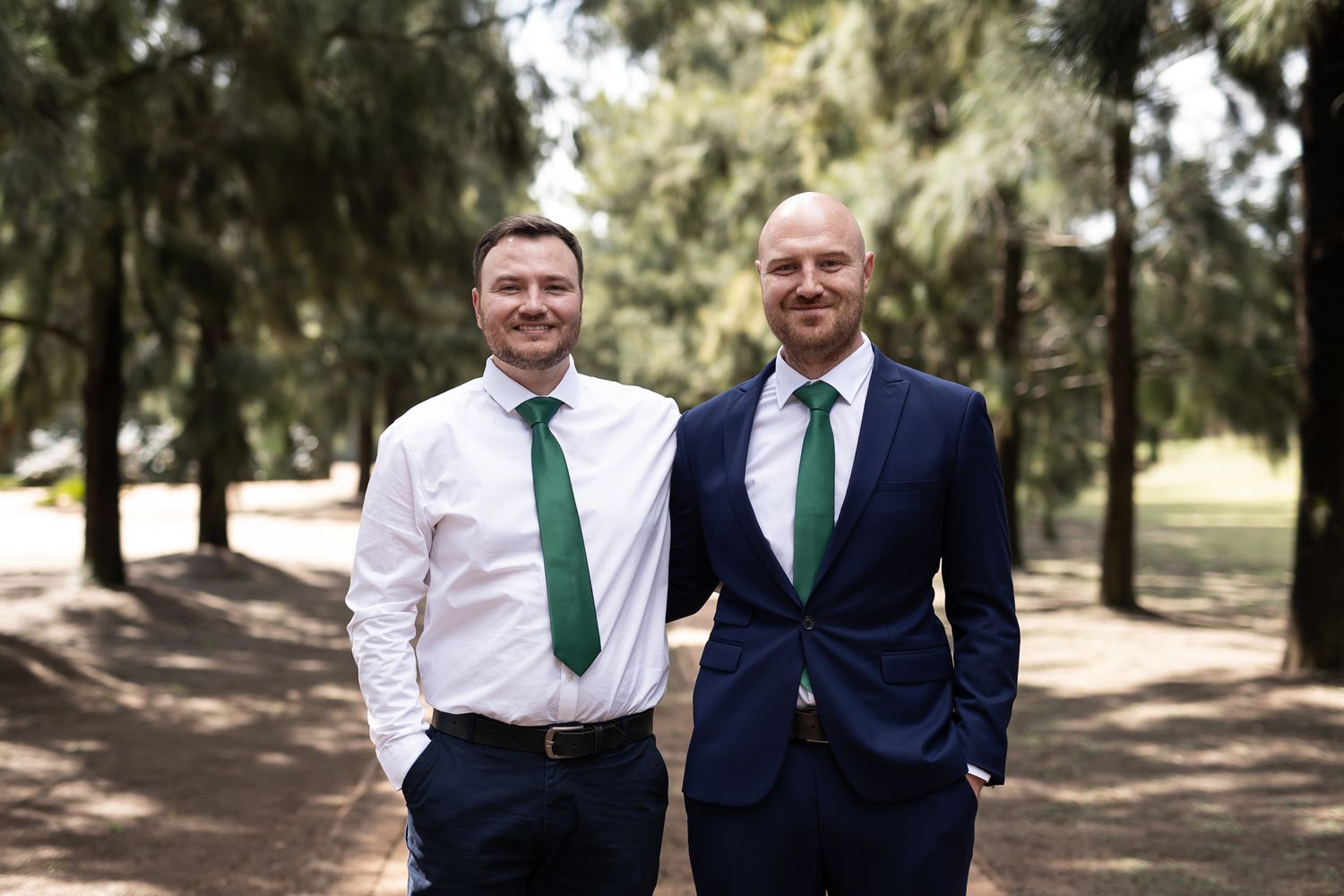 Two men in coordinated formal wear with green ties smiling in wooded setting.