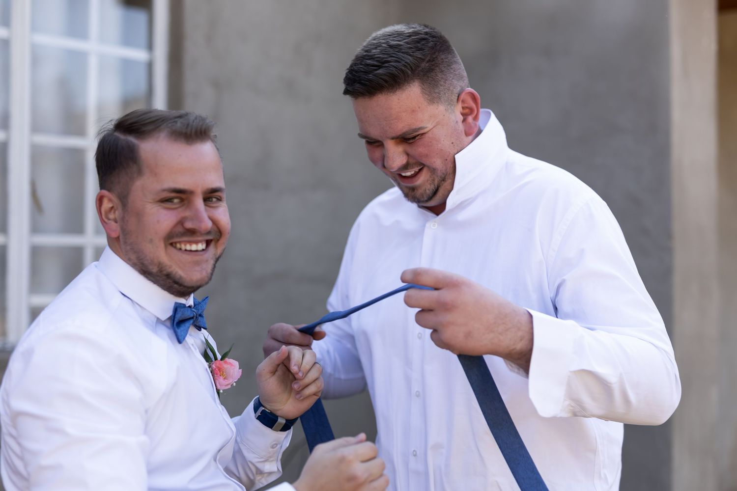 Two people in white dress shirts share a lighthearted moment while adjusting a blue tie before a formal event.