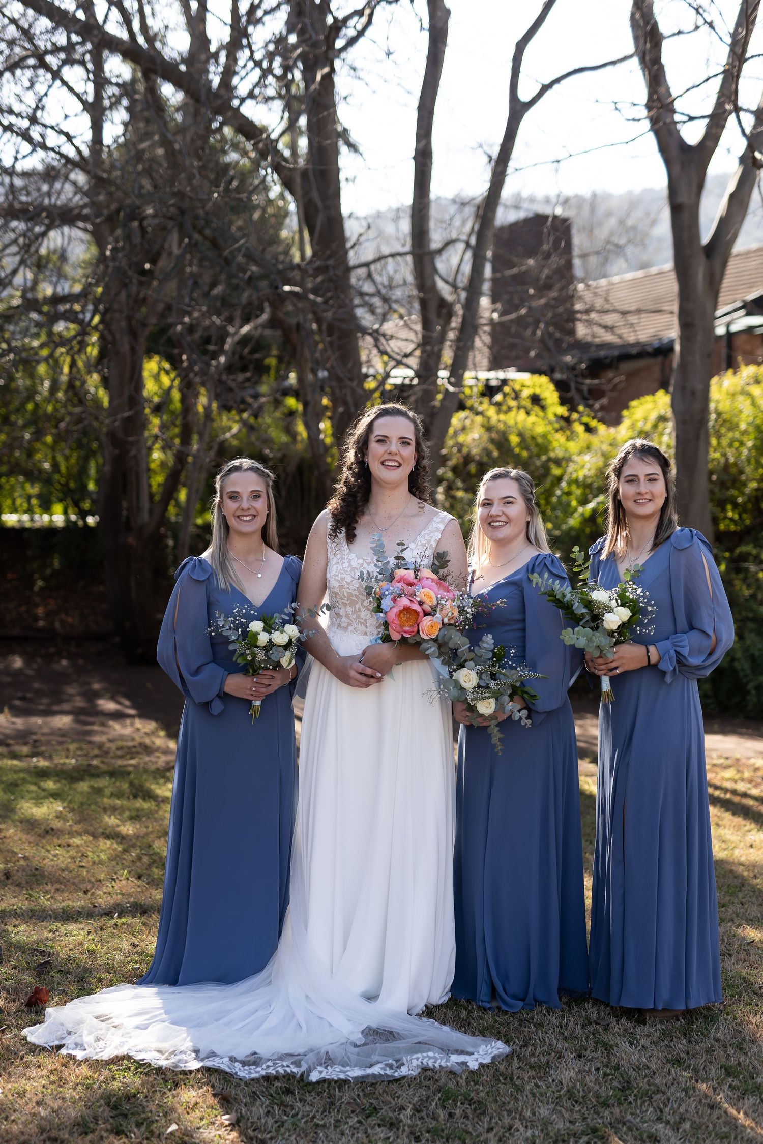 A bridal party in navy blue bridesmaid dresses and white wedding gown poses outdoors in natural garden setting.