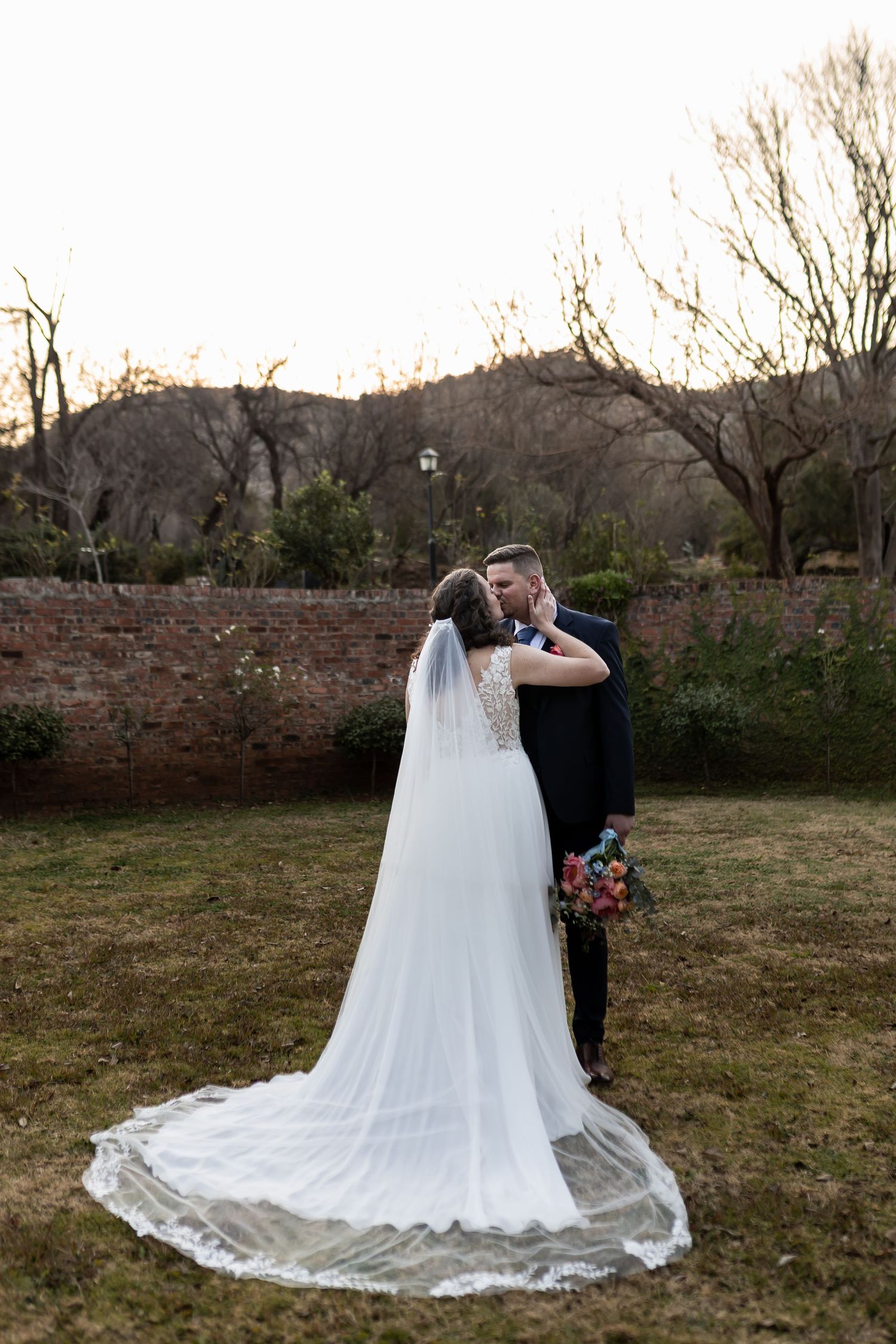 Beautiful wedding couple embraces in front of a rustic brick wall at dusk with bride wearing elegant lace-trimmed veil and gown.