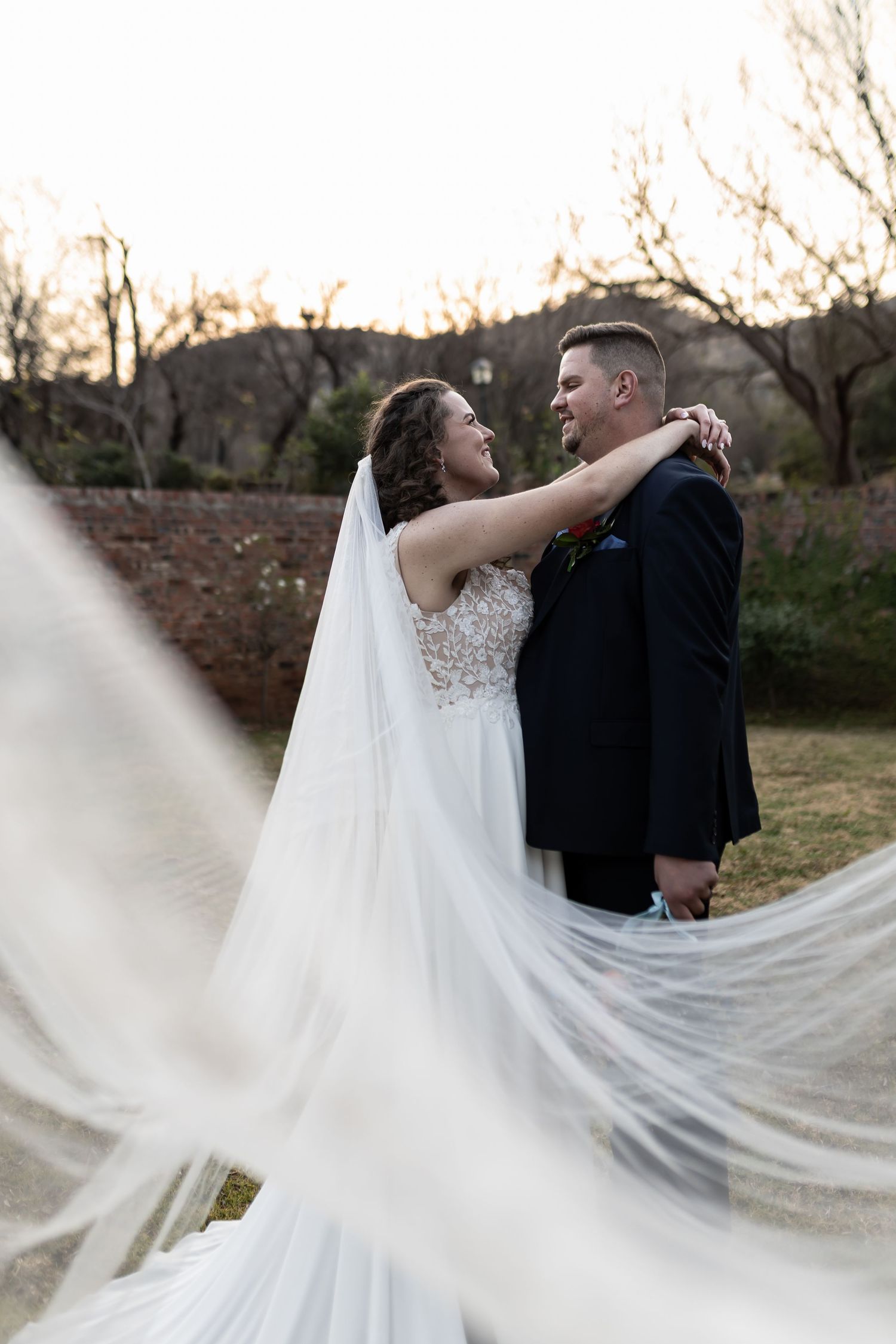 A romantic wedding couple dances in an outdoor setting as the bride's flowing white veil gracefully moves in the wind.