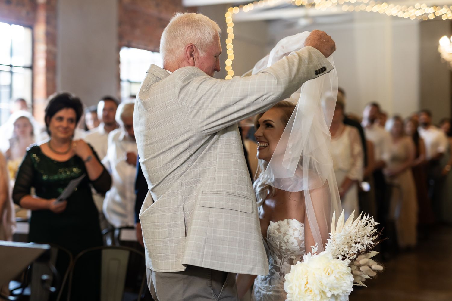 Tender moment during wedding ceremony with brick walls and string lights in background.