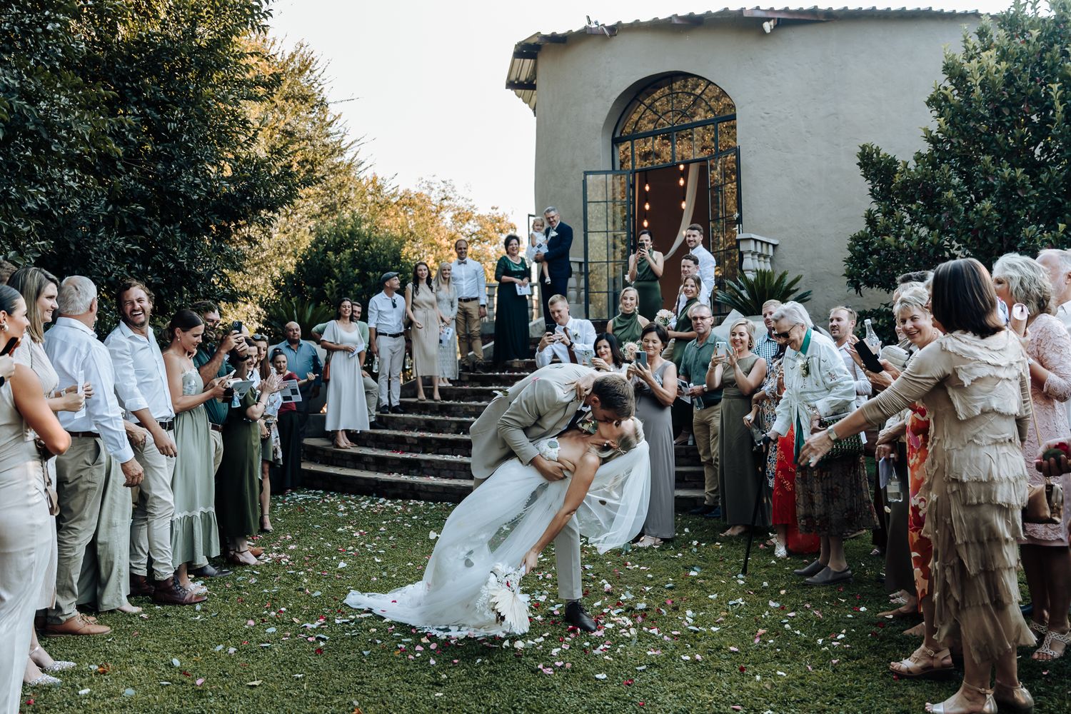 Newlyweds share a romantic dip kiss while guests celebrate with confetti outside a church.