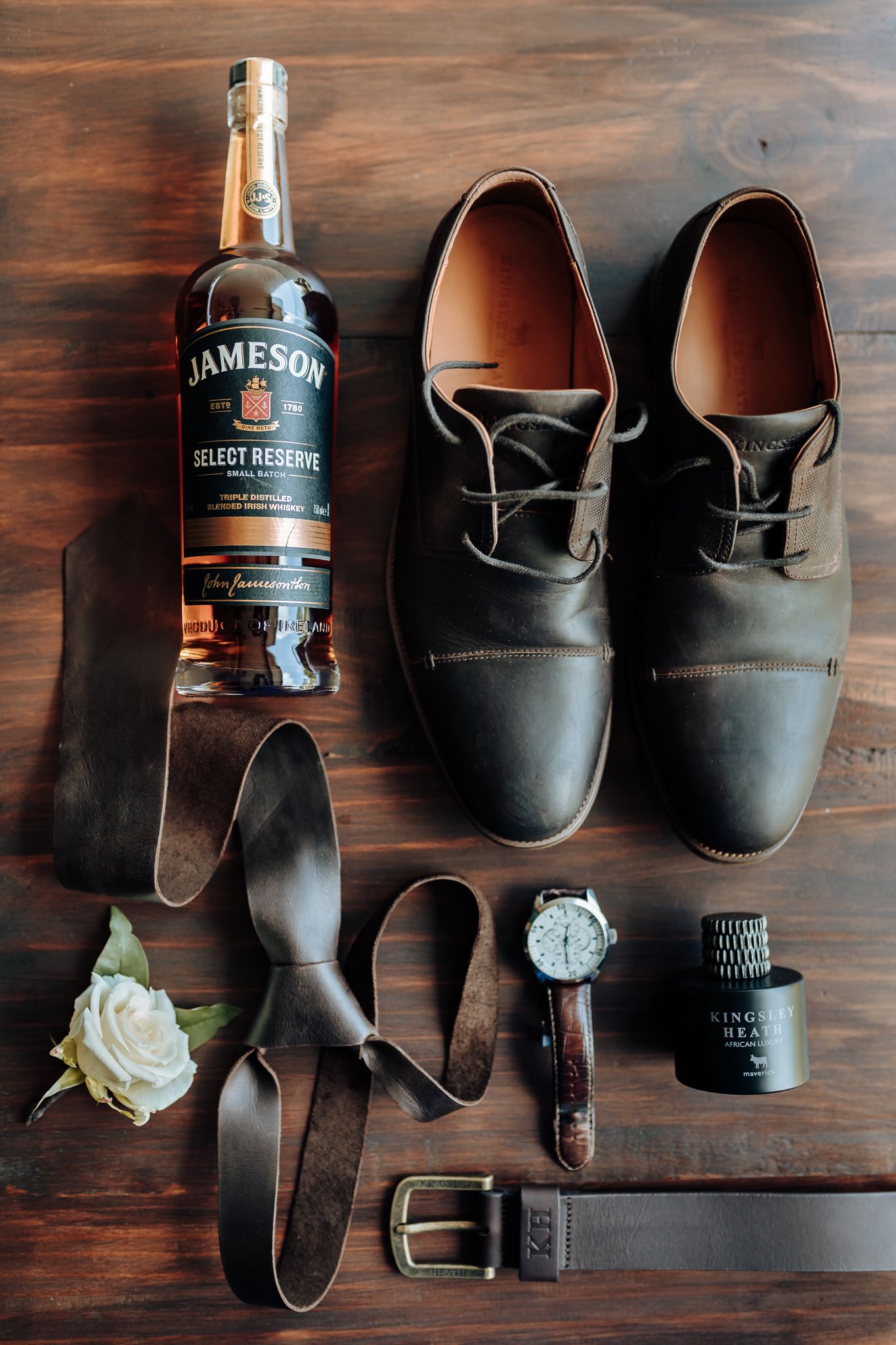 Groom's accessories including Jameson whiskey, dress shoes, watch and belt laid on wooden surface.