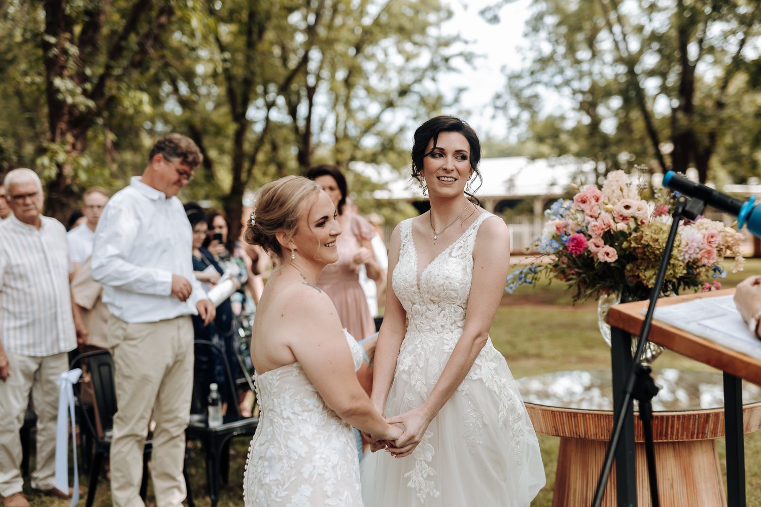 Two people in white wedding dresses share a joyful moment during outdoor ceremony.