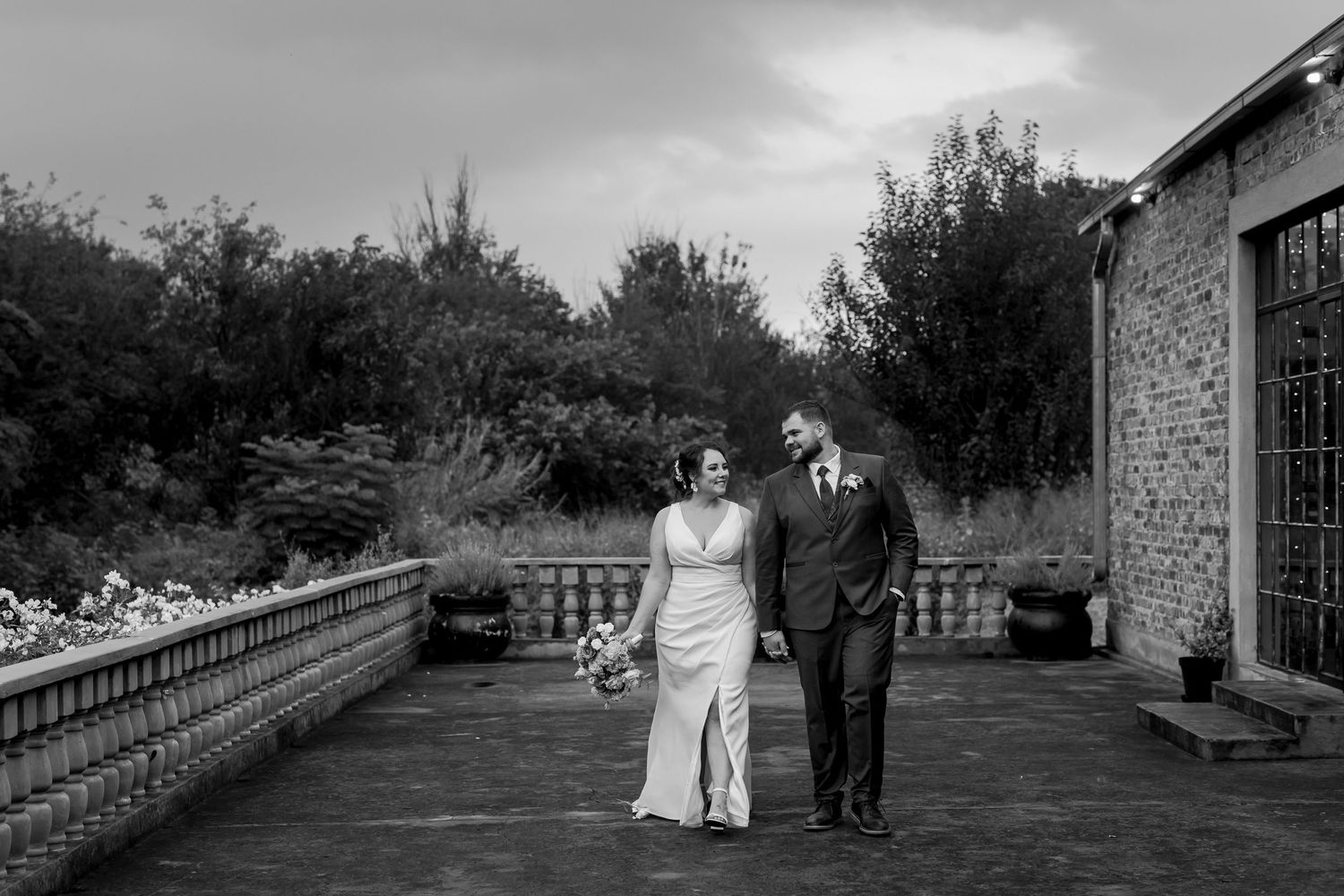 Black and white photo of couple walking along stone balustrade at wedding venue.