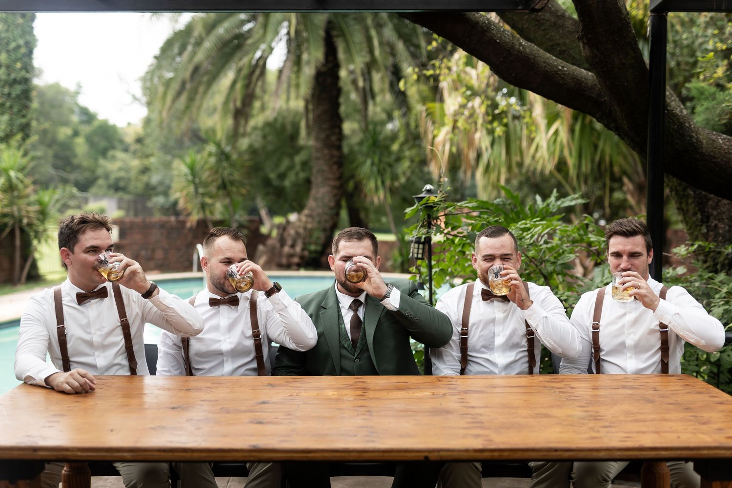Groomsmen taking shots together at a wooden table in a tropical garden setting.