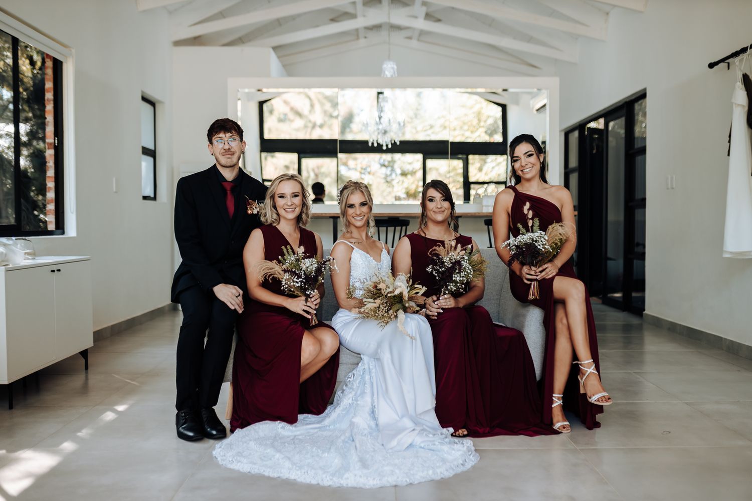 Wedding party poses together in burgundy dresses and formal attire in elegant white venue.
