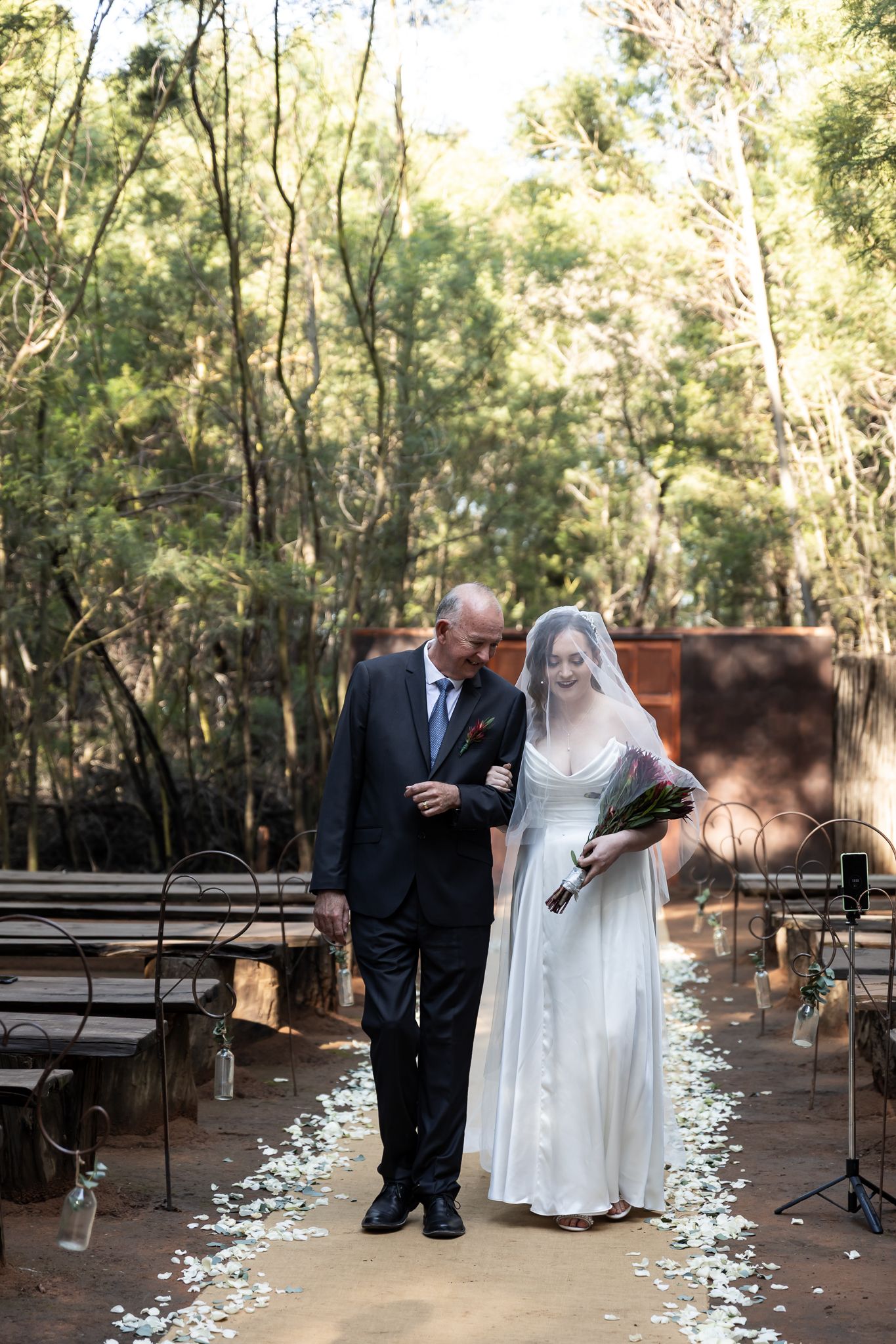 A traditional wedding ceremony processional along a petal-strewn aisle in a wooded outdoor setting.