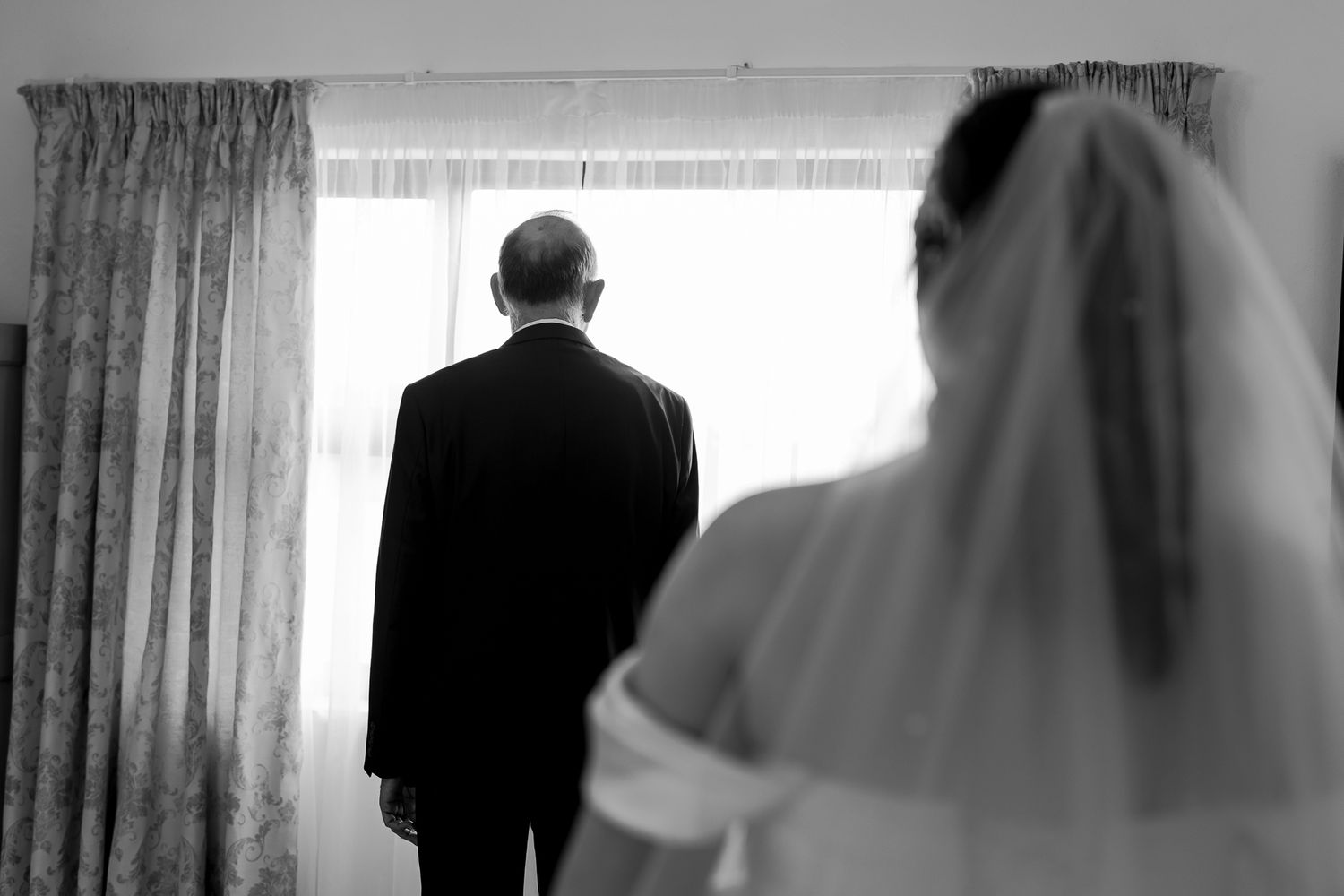 Black and white silhouette of figures by window in wedding attire.