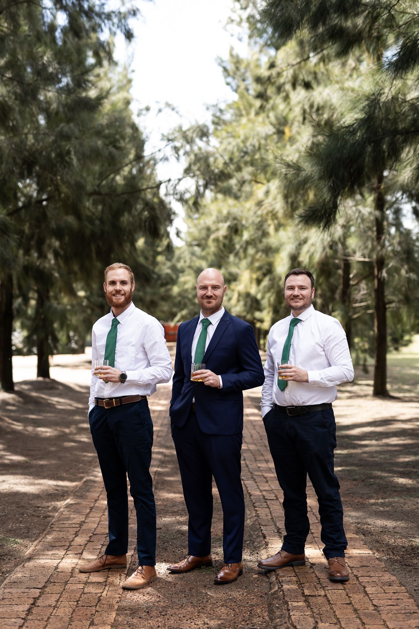 Three men in formal attire with green ties standing on brick pathway among pine trees.