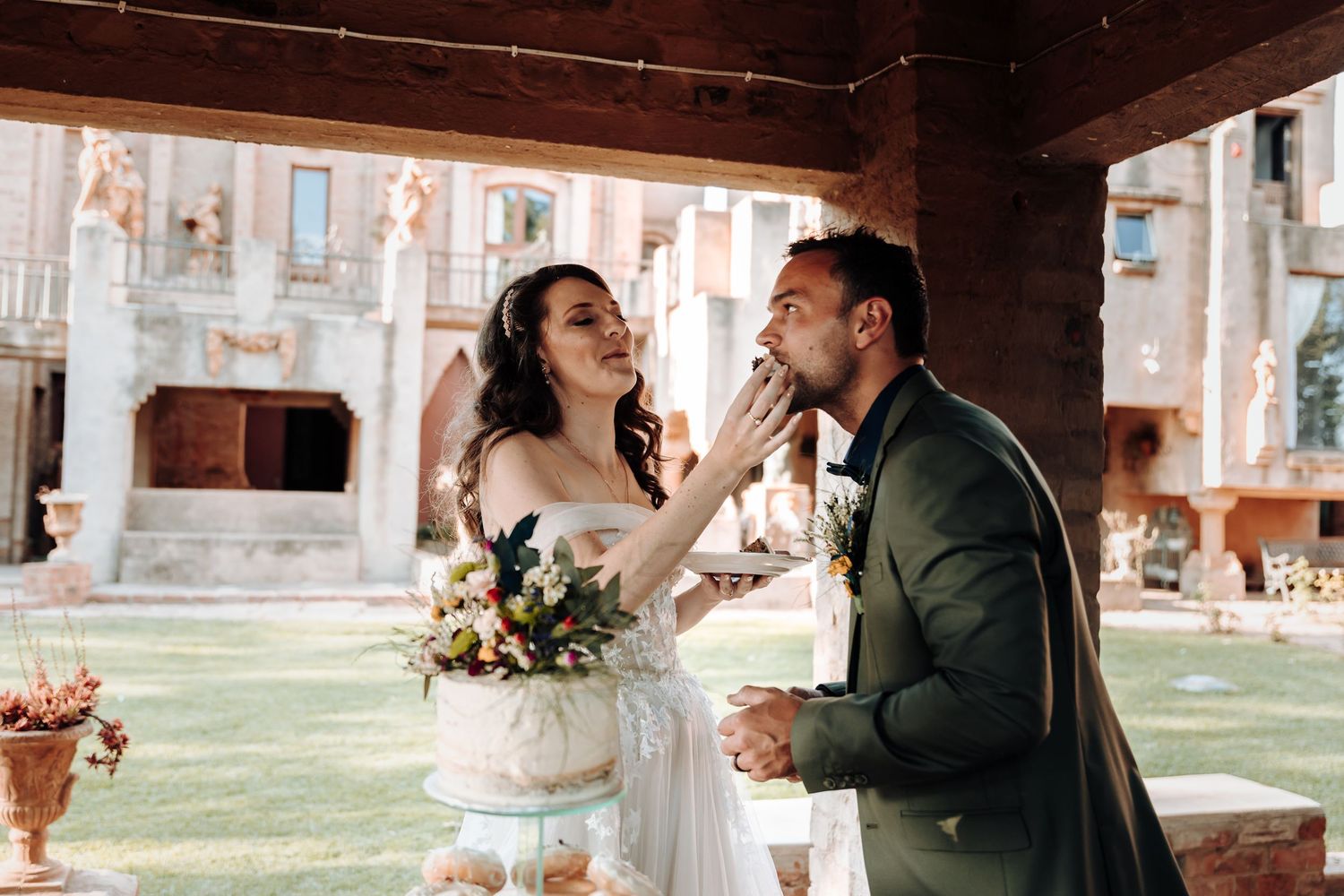 Couple laughs while sharing cake at their wedding reception in an outdoor courtyard.