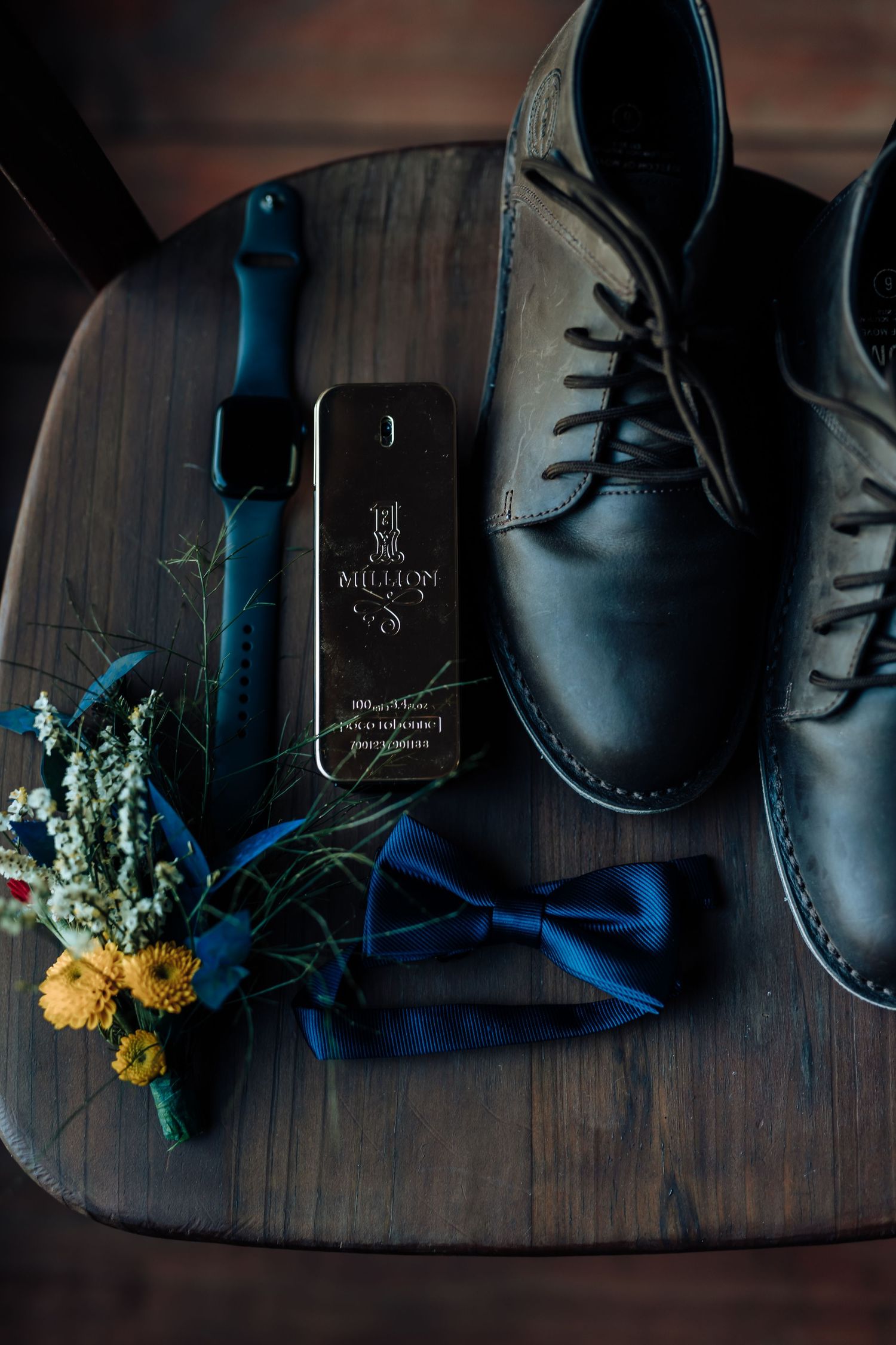 Groom's accessories featuring black dress shoes, blue bowtie, and boutonniere on wooden surface.
