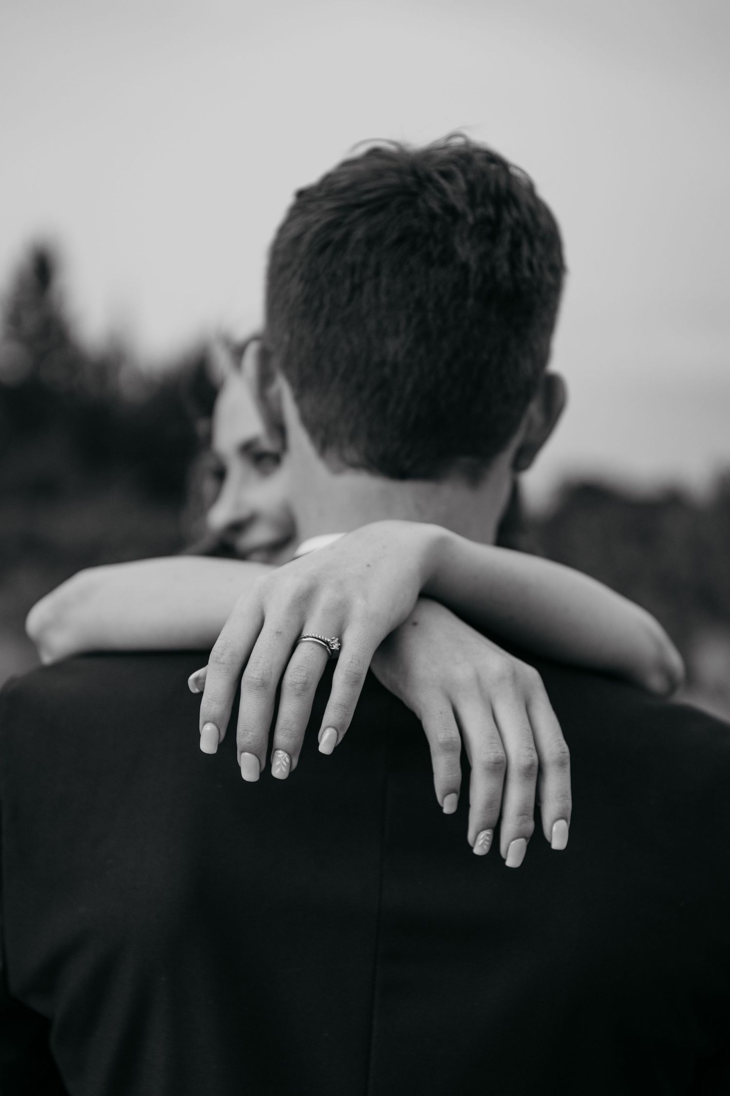 Black and white photo showing bride's hands with ring embracing groom from behind.