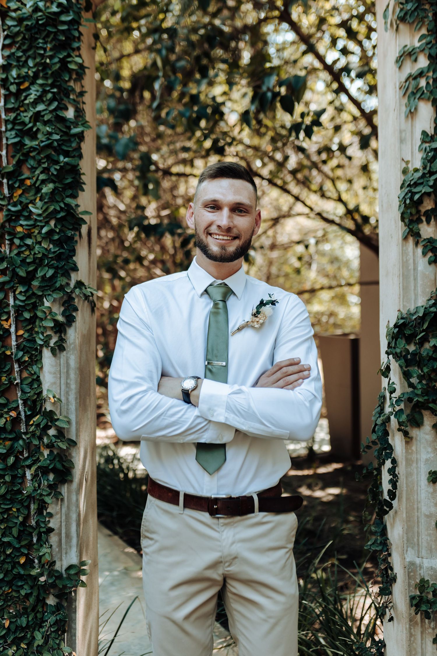 Person in white shirt and sage tie poses confidently with arms crossed in garden archway.