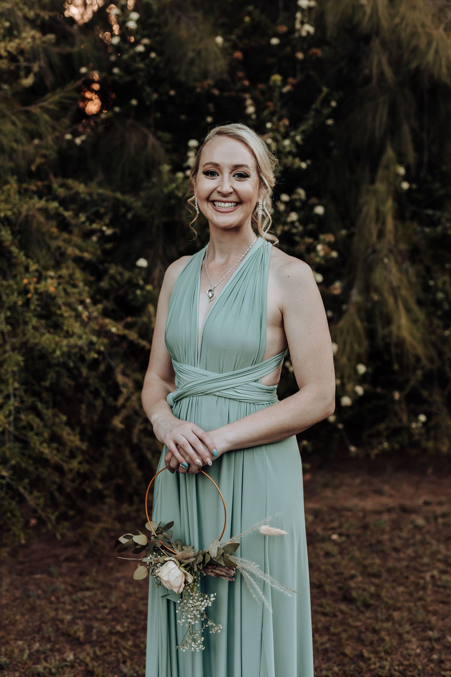 Woman in flowing mint green bridesmaid dress poses with bouquet at outdoor wedding venue.