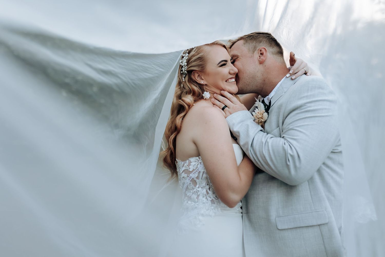 Tender wedding moment with dramatic flowing veil and light gray suit against white background.