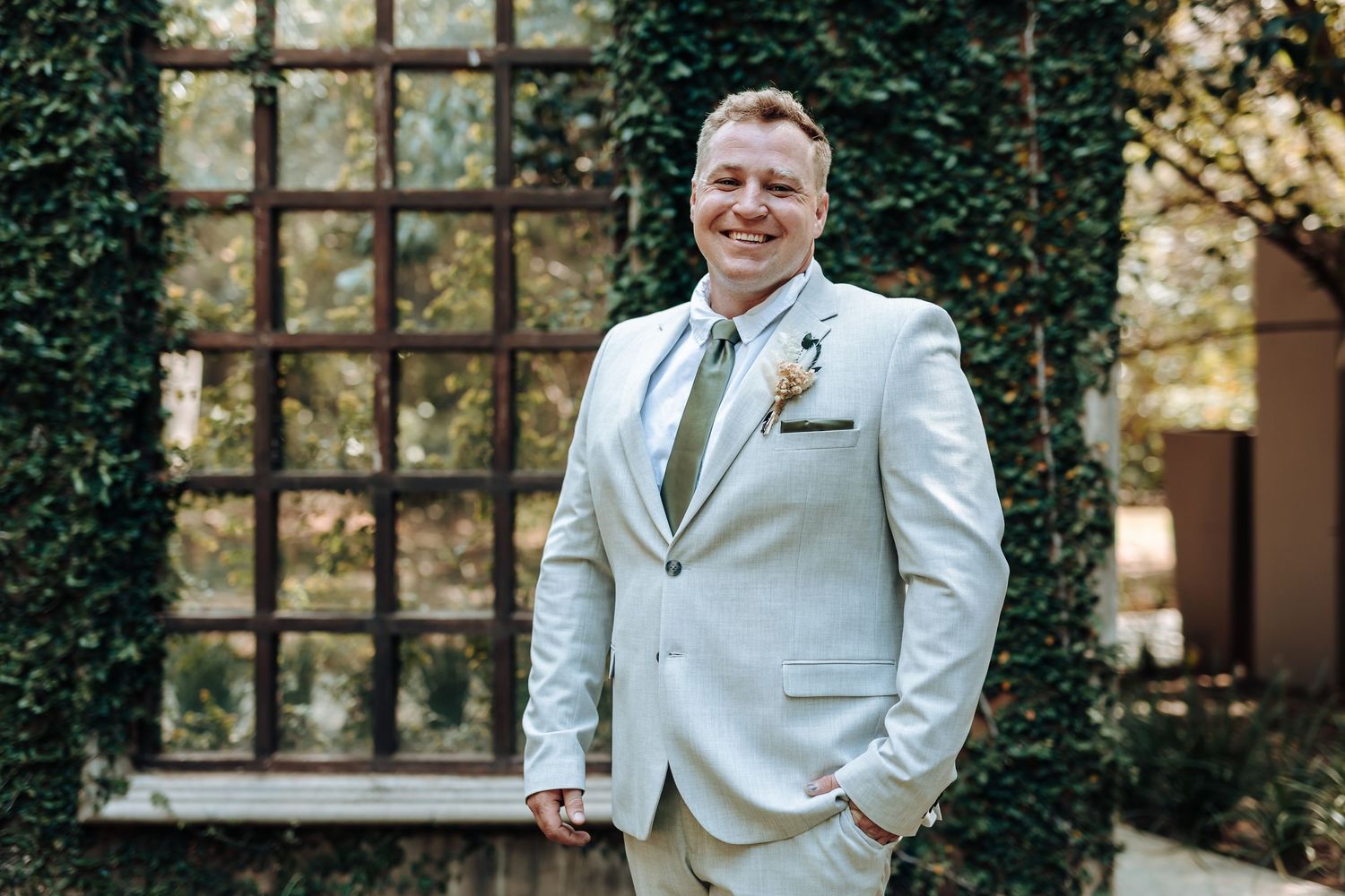 Groom in light gray suit with sage tie poses confidently against garden wall backdrop.