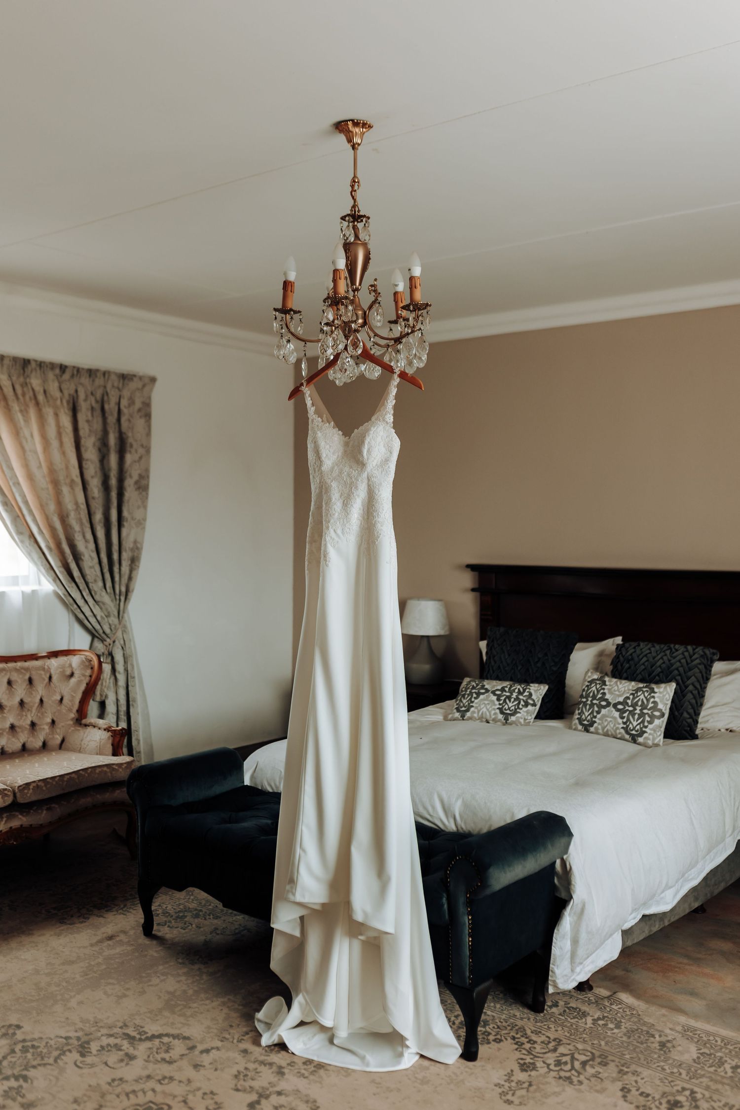 Elegant white wedding dress hanging from crystal chandelier in luxurious bedroom.