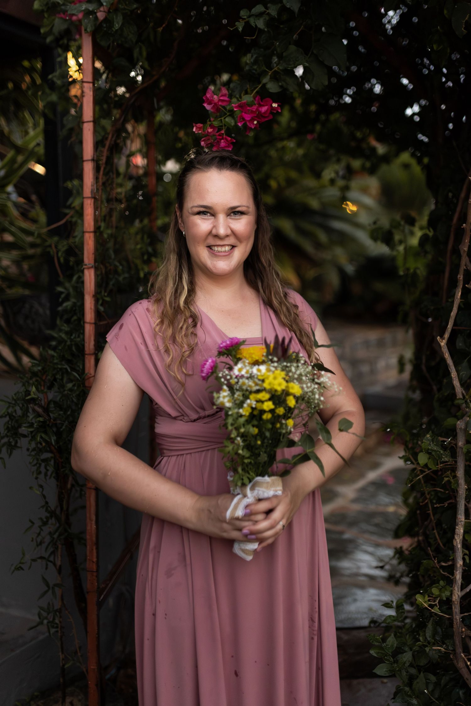 Bridesmaid in dusty rose dress holding cheerful bouquet of yellow and purple wildflowers.