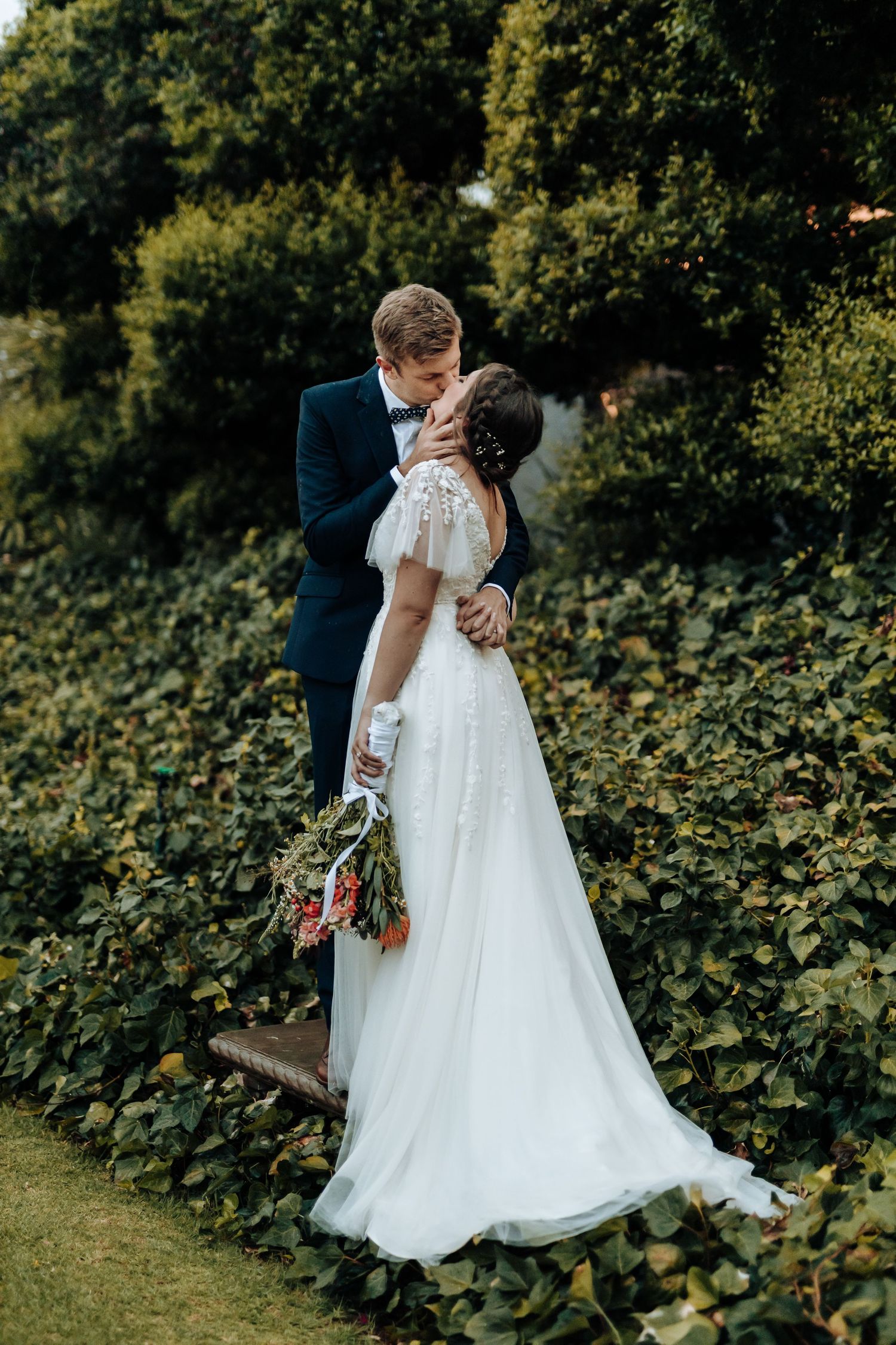 Romantic wedding couple embracing in a lush garden setting with the bride holding a vibrant bouquet.