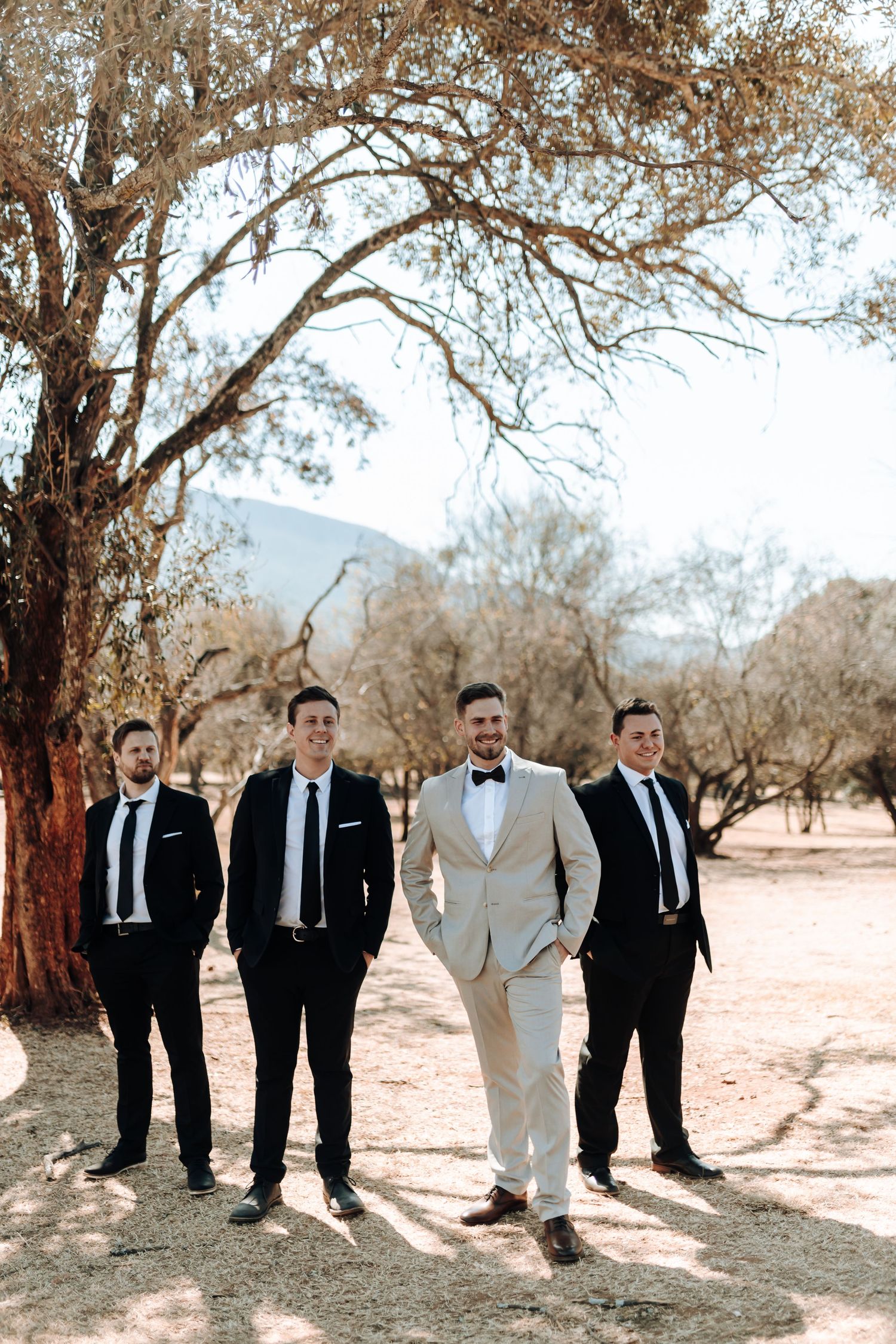 Formal wedding party poses together under trees with central figure in light colored suit.