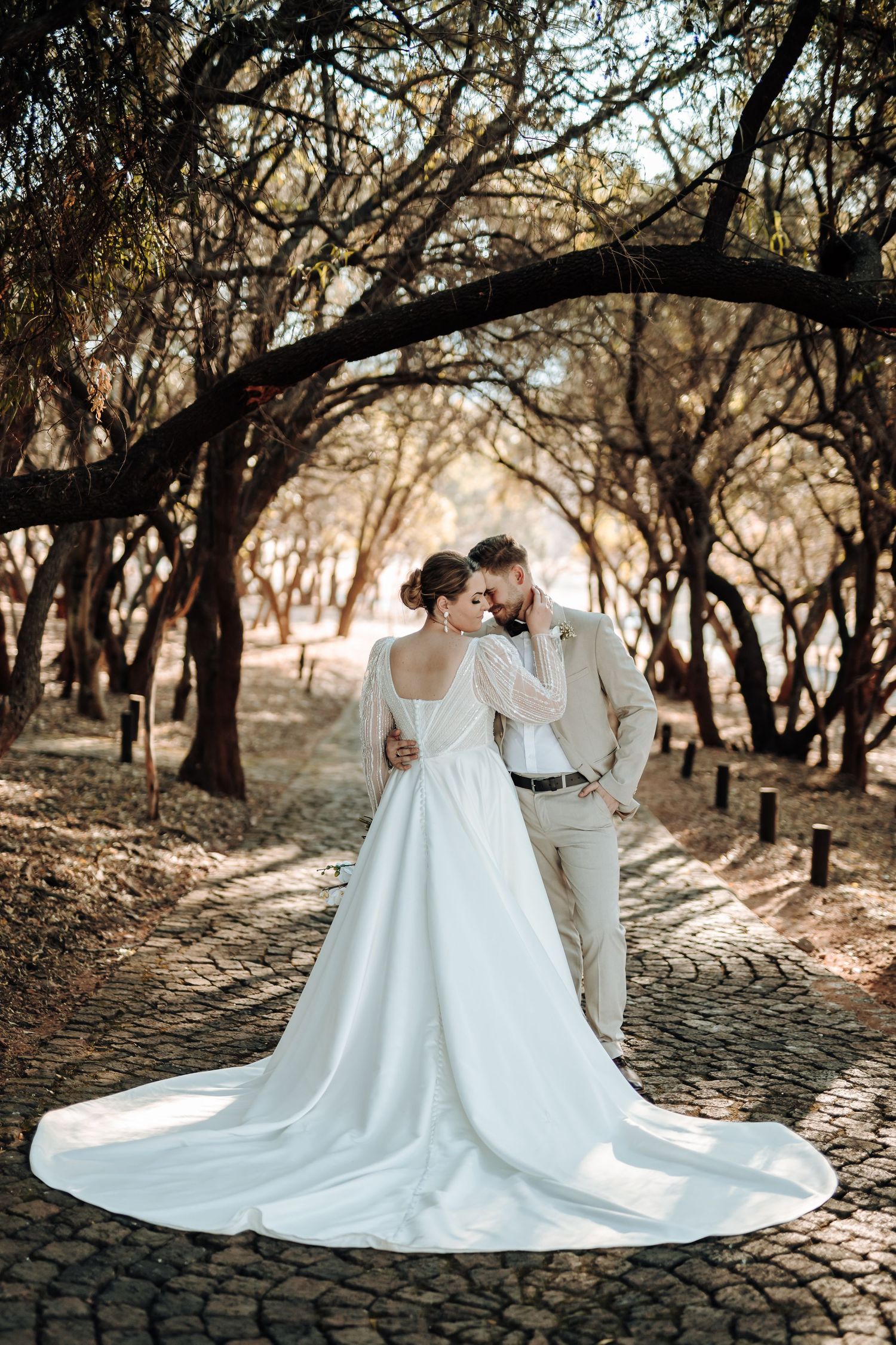 Bride in flowing white gown and groom share intimate moment on cobblestone forest path.