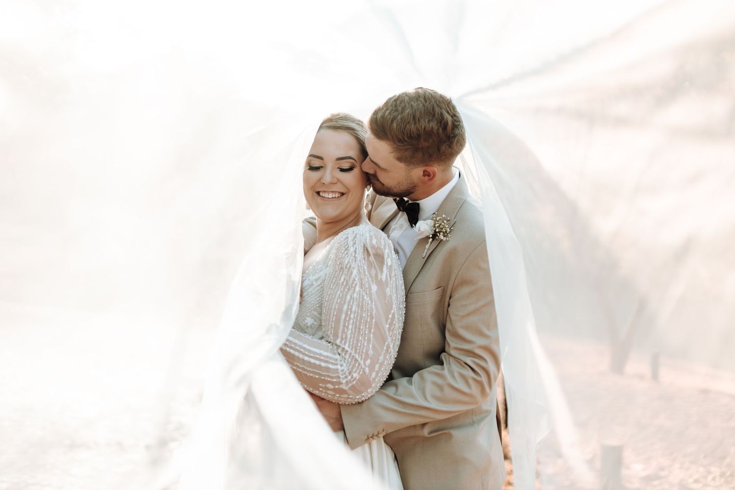 Intimate wedding portrait with bride in lace dress and groom in tan suit against ethereal backdrop.