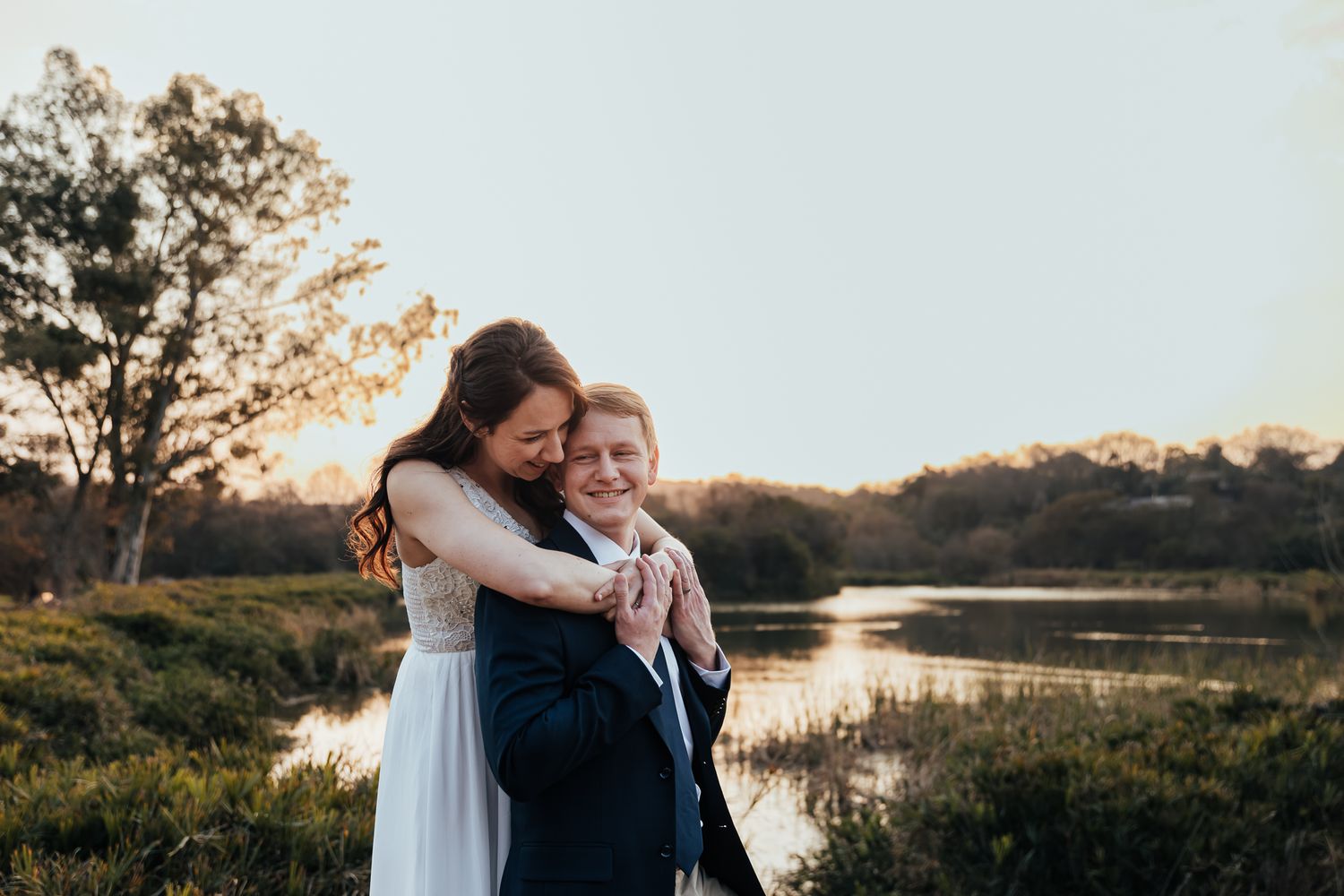 Couple embraces by waterfront during sunset with trees and marsh in background.