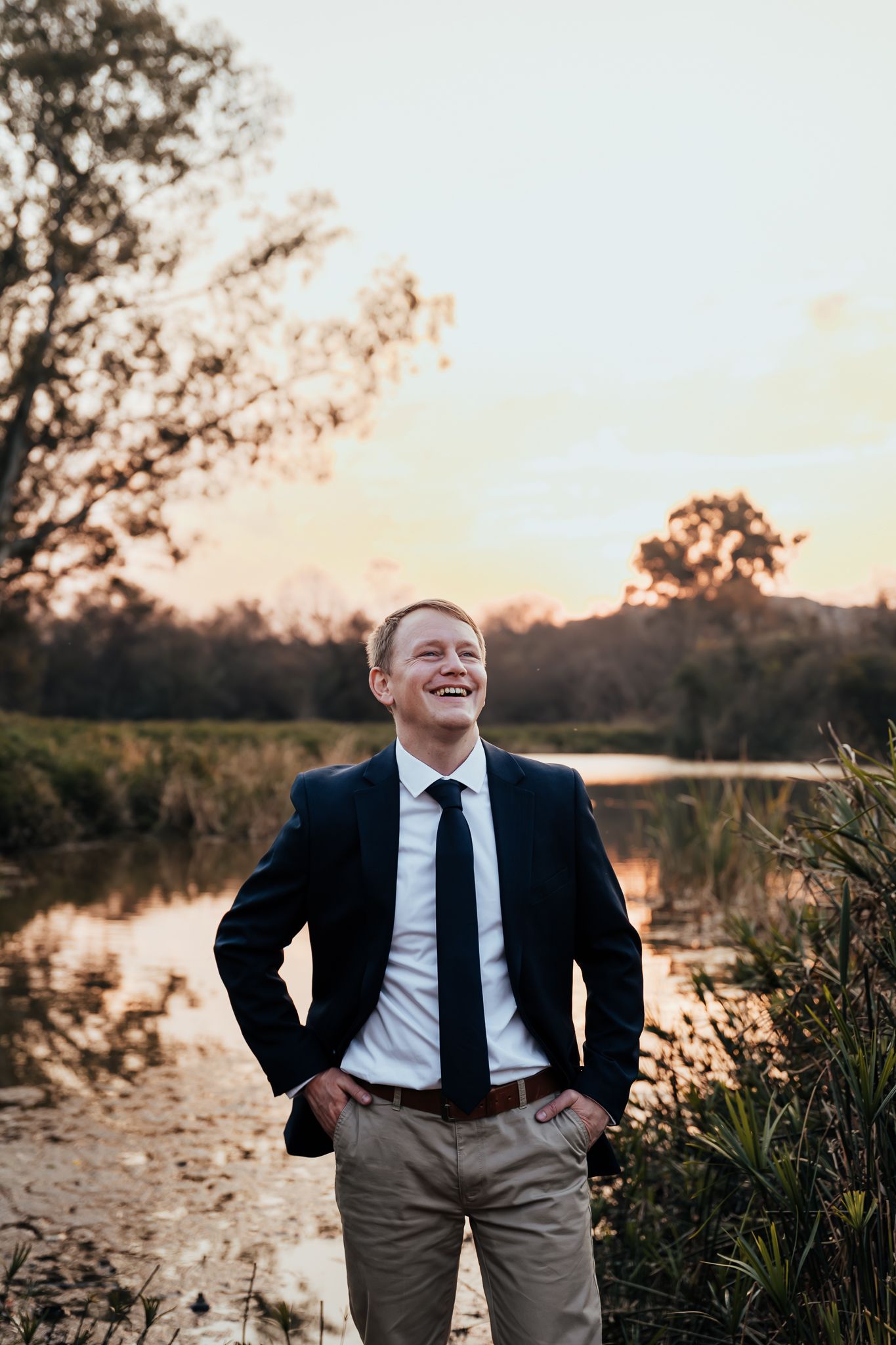 A groom smiles while standing near a scenic pond during sunset with trees reflected in the water.