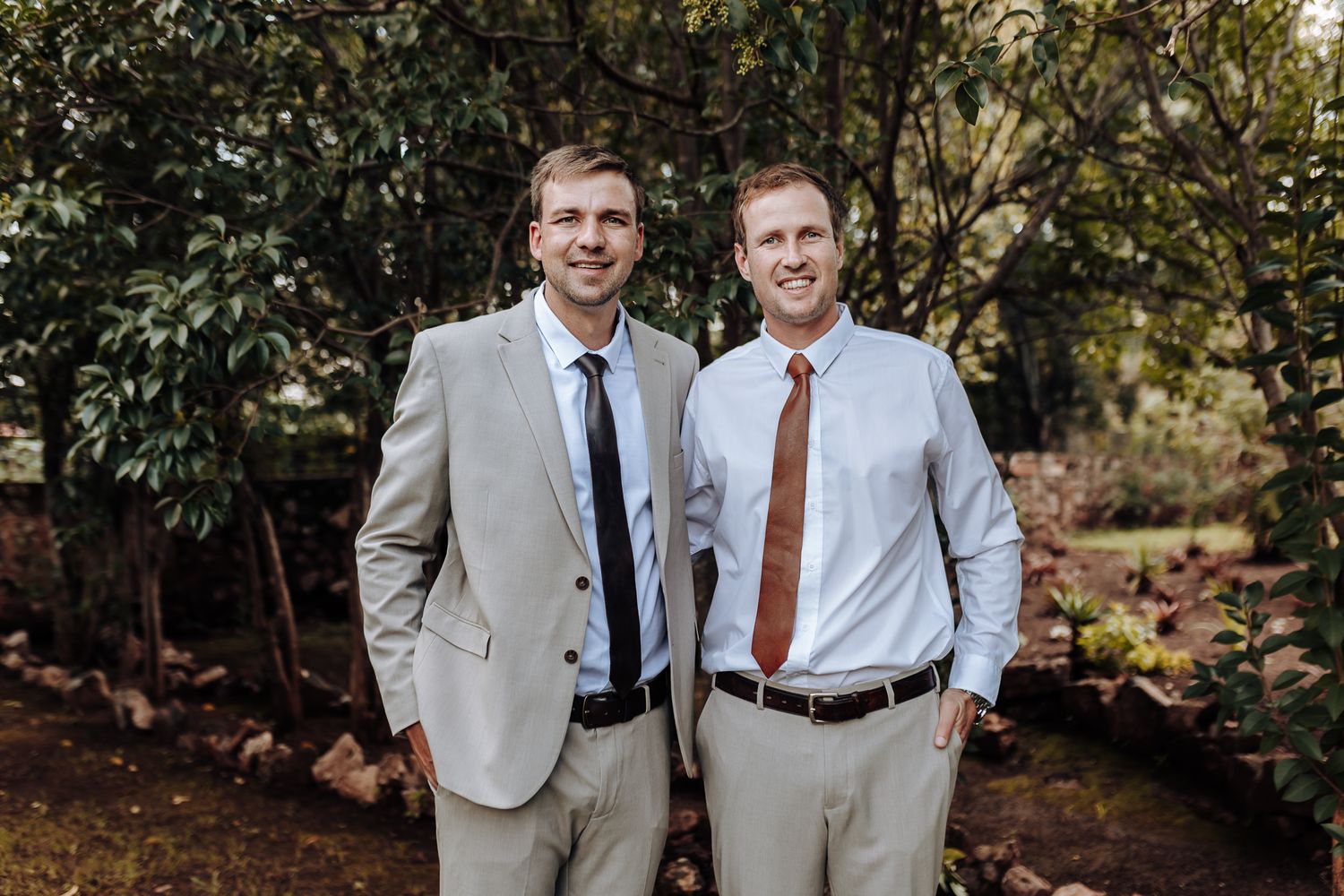 Two men in light suits and contrasting ties pose together against foliage.