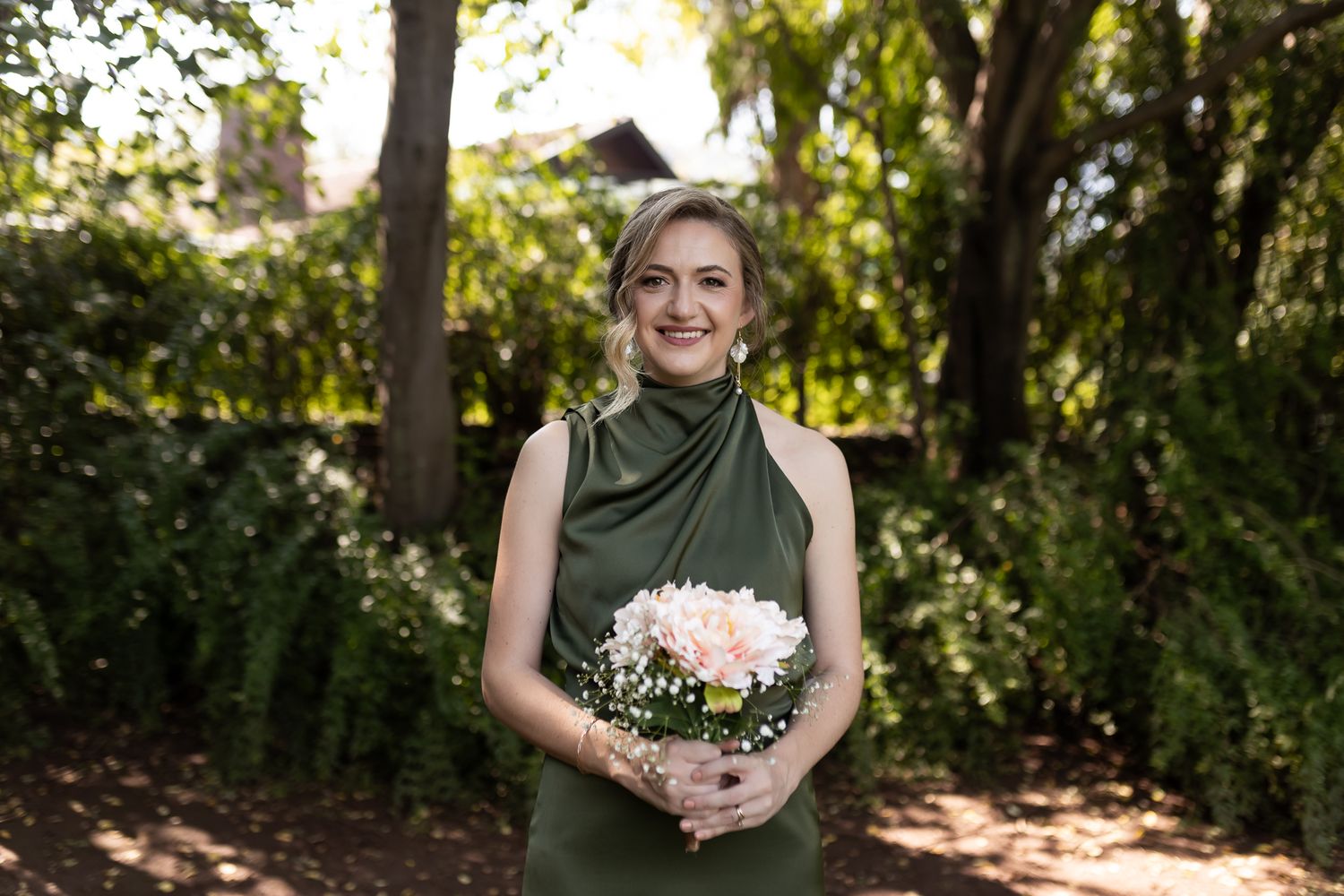 Bridesmaid in elegant olive green halter dress holds white bouquet in garden setting.