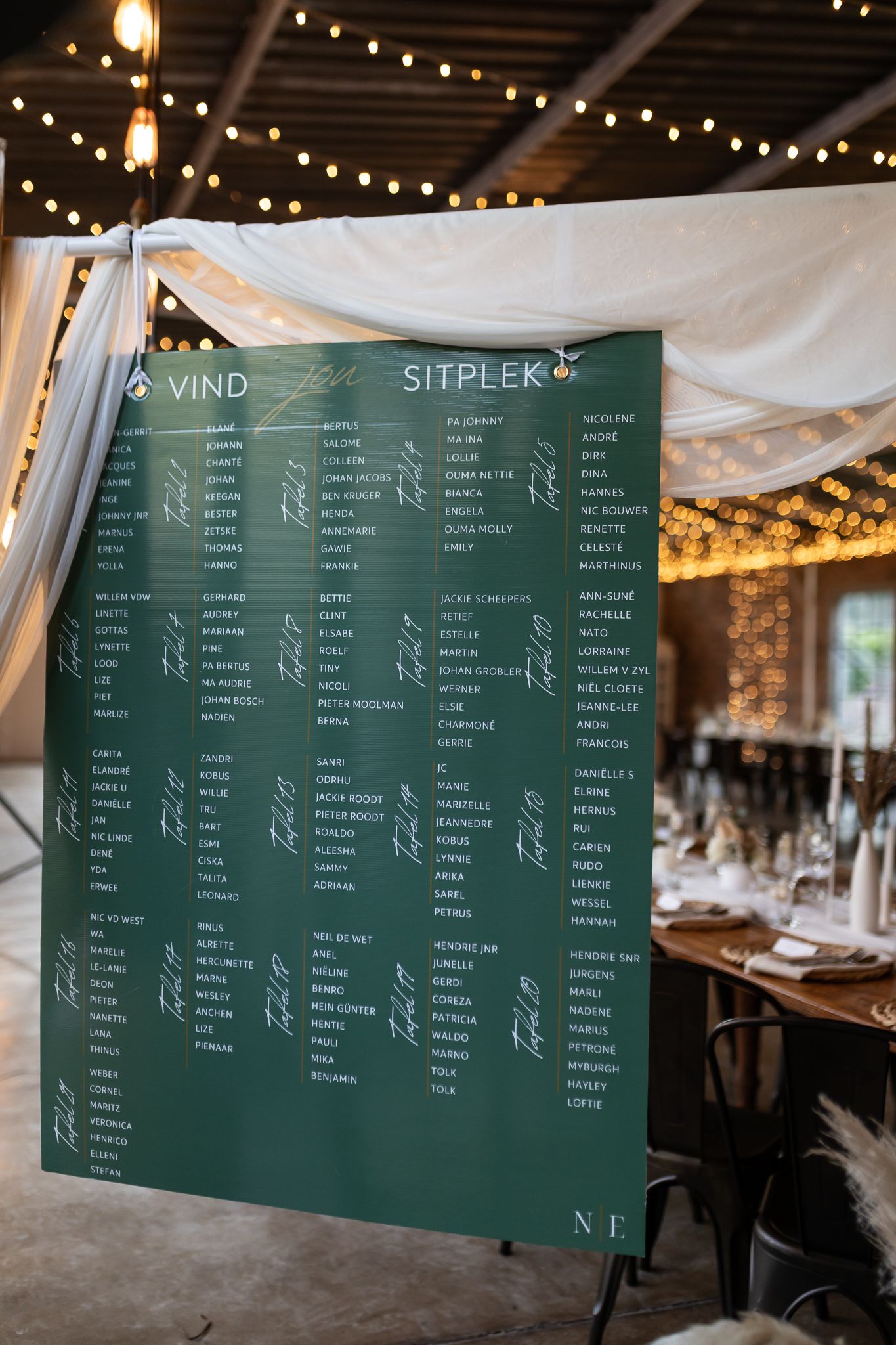 Green wedding seating chart display with string lights and white fabric draping in reception venue.