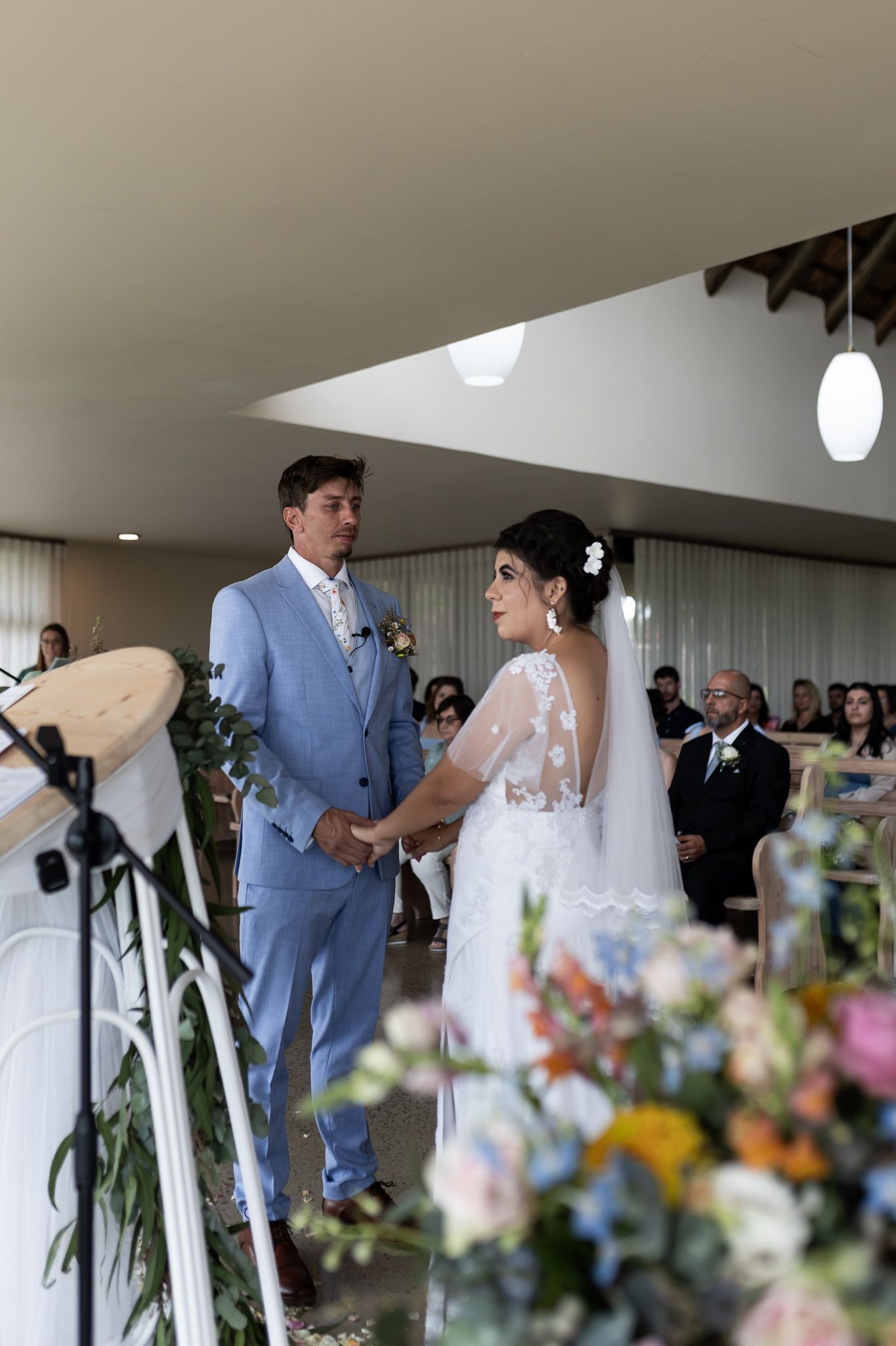 A couple holds hands during their wedding ceremony surrounded by elegant floral arrangements.