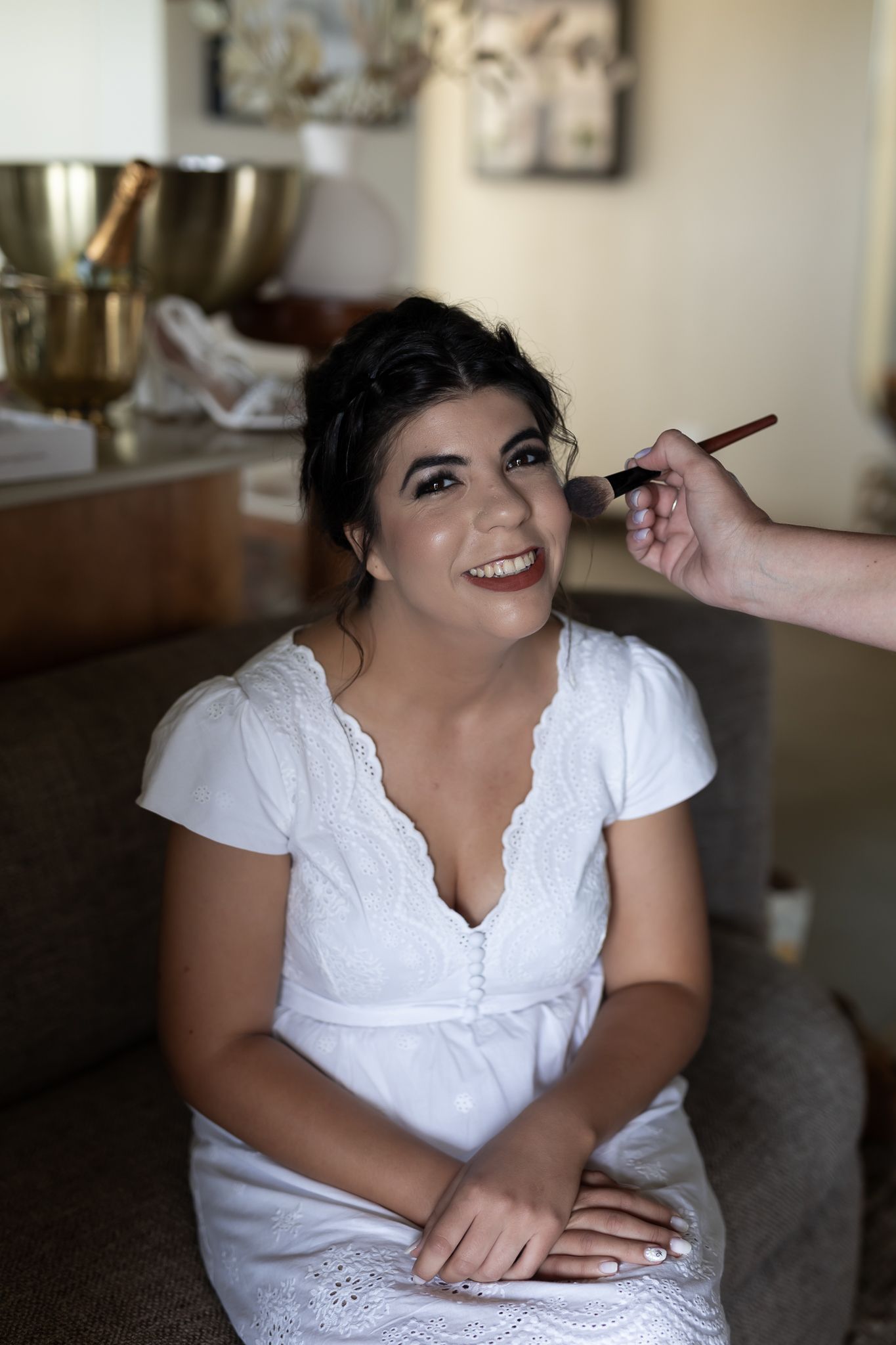 A person in a white dress getting makeup applied while sitting on a couch.