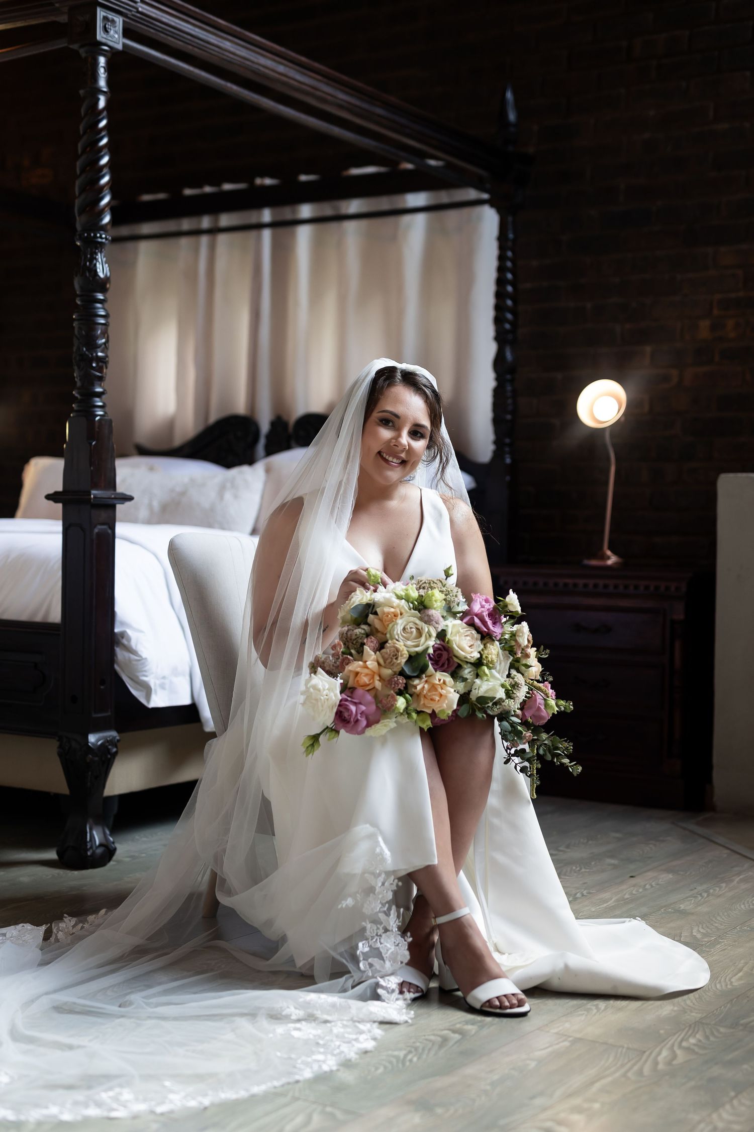 Bride in white gown sits with flowing veil and bouquet against dark wood four-poster bed.