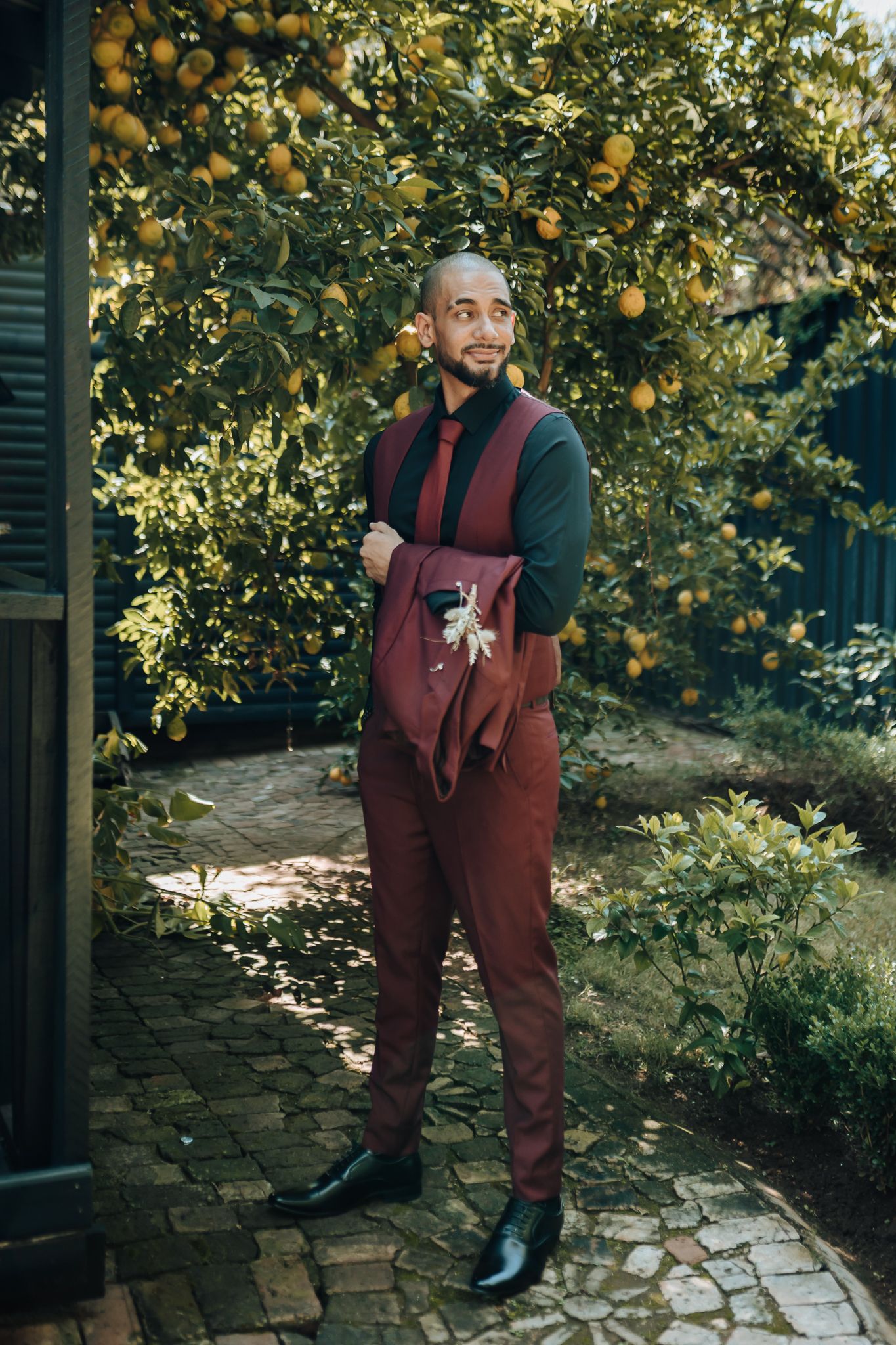 Stylish groom wearing burgundy overalls and black shirt poses in a citrus garden.
