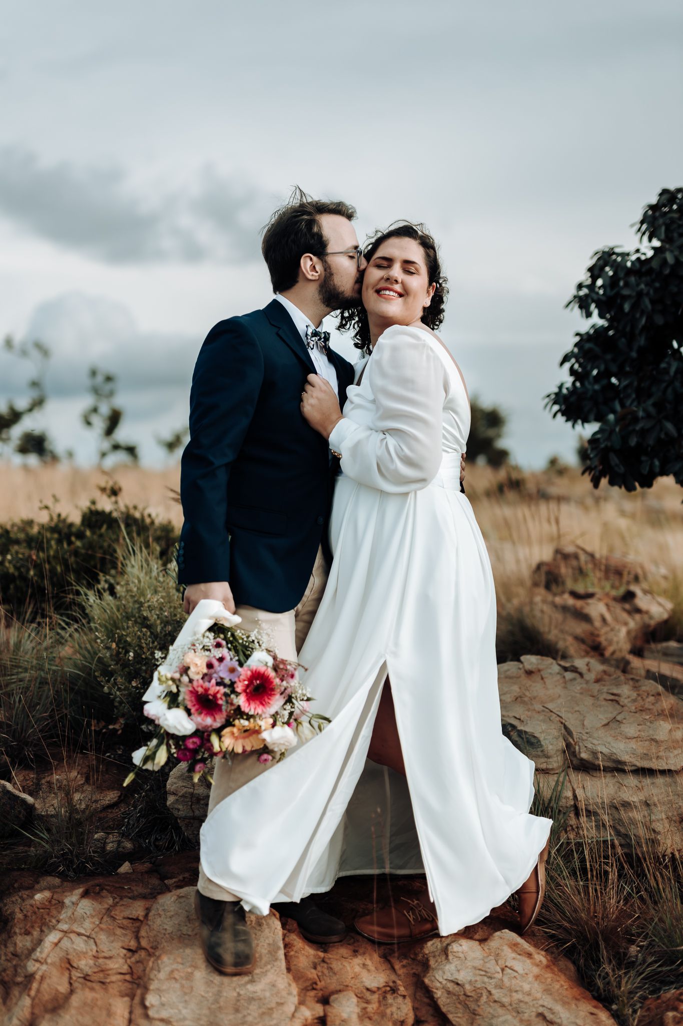 A romantic moment captured between newlyweds on a rocky hillside with a colorful bouquet.