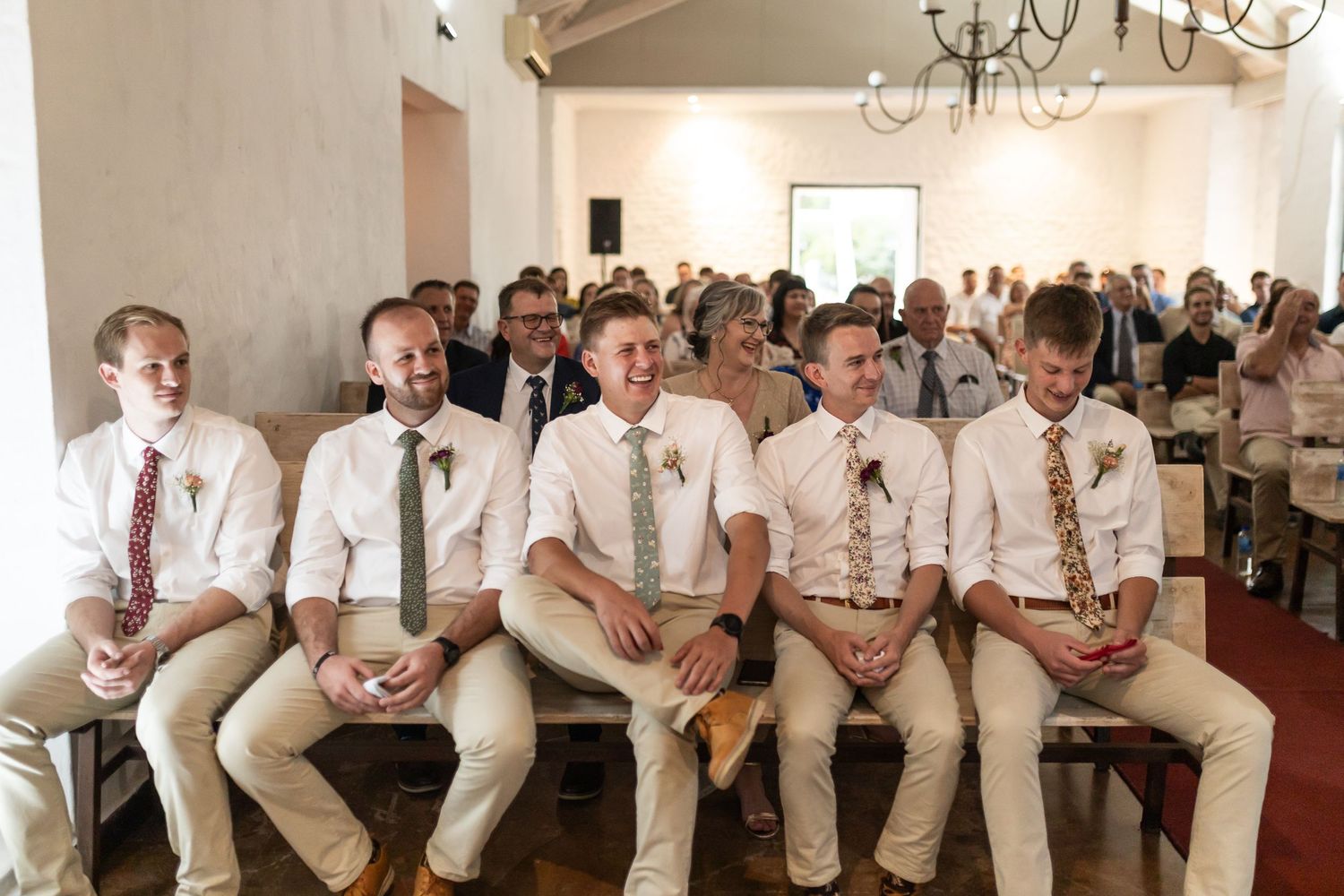 Groomsmen wearing khaki pants and white shirts sit in a row during a wedding ceremony.