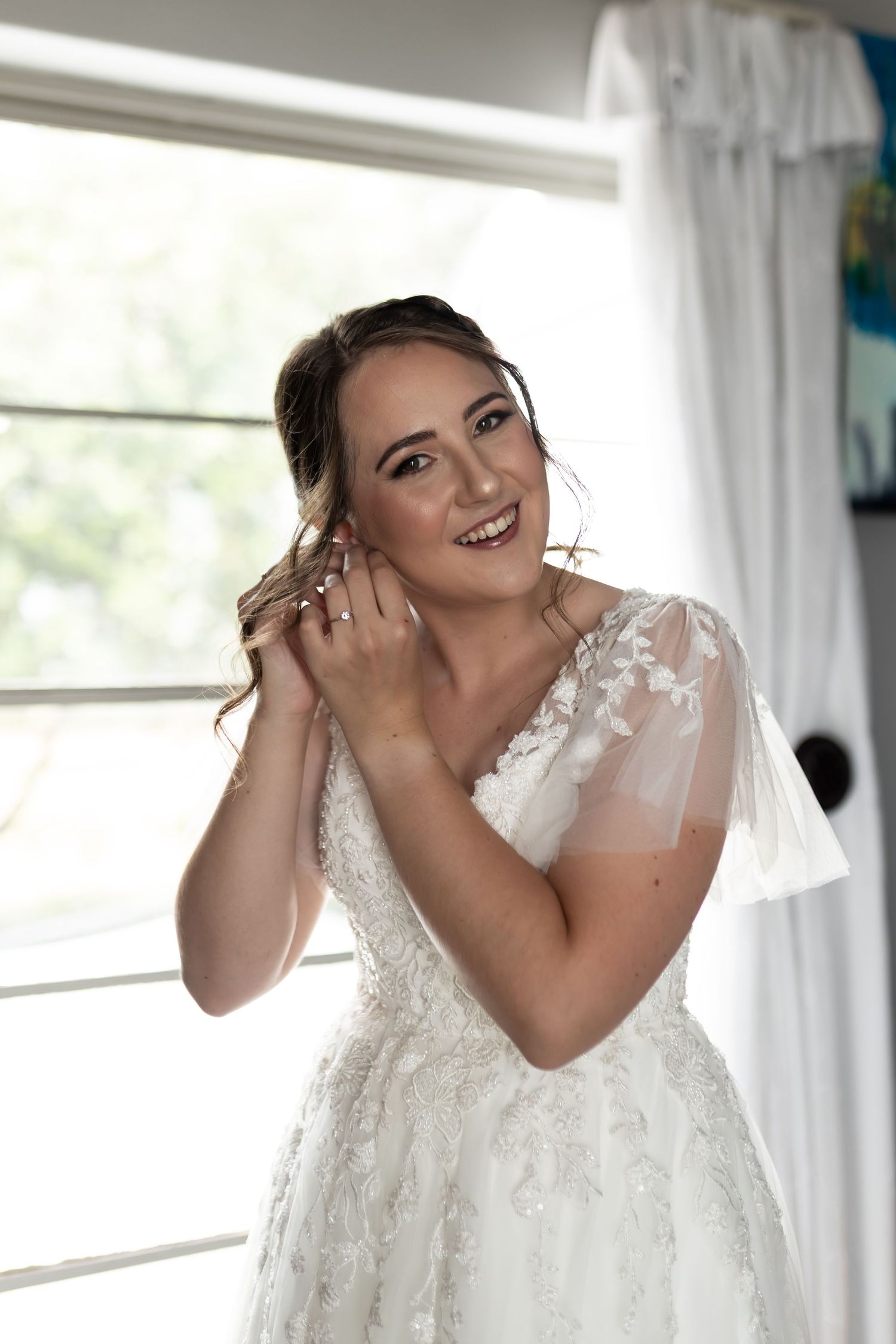 A person in a white wedding dress smiles while putting on earrings near a window.