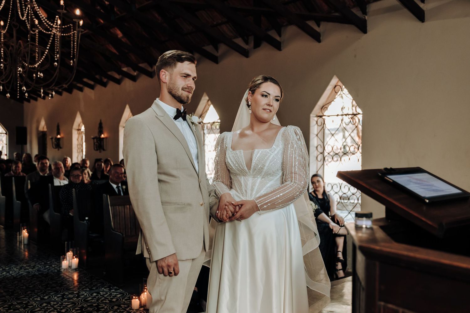 A couple holds hands during their wedding ceremony in a chapel with gothic architecture.