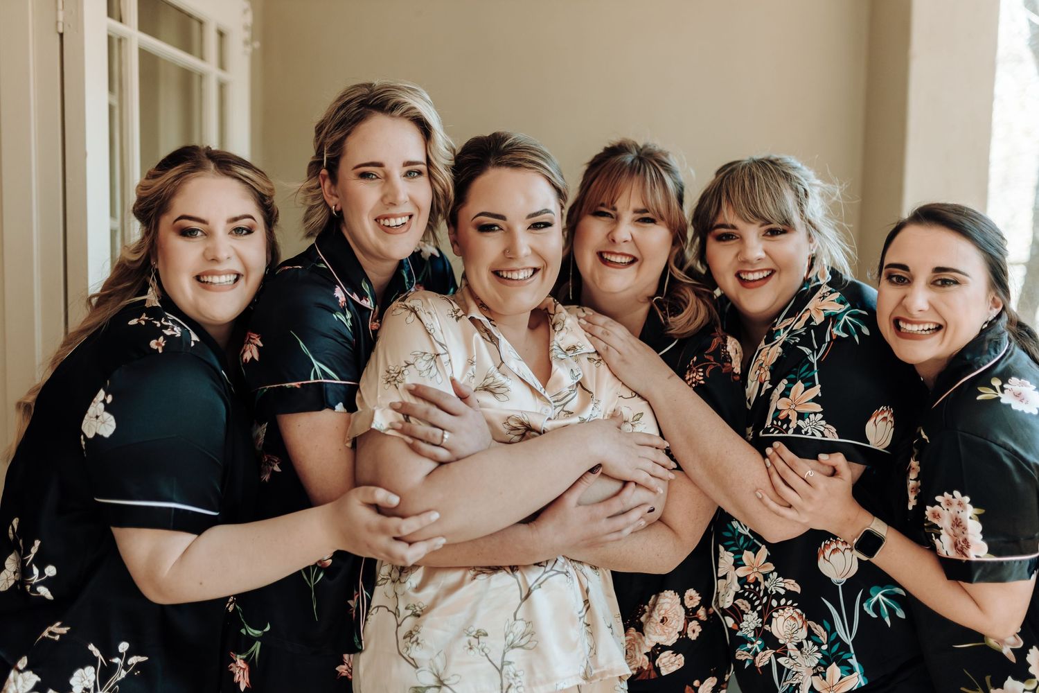 Bridesmaids in black floral robes embracing the bride in white during pre-wedding celebrations.