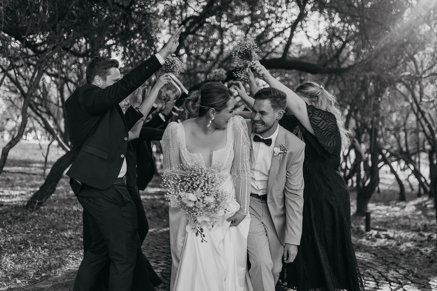 Black and white photo of wedding party creating an arch over couple in woods.